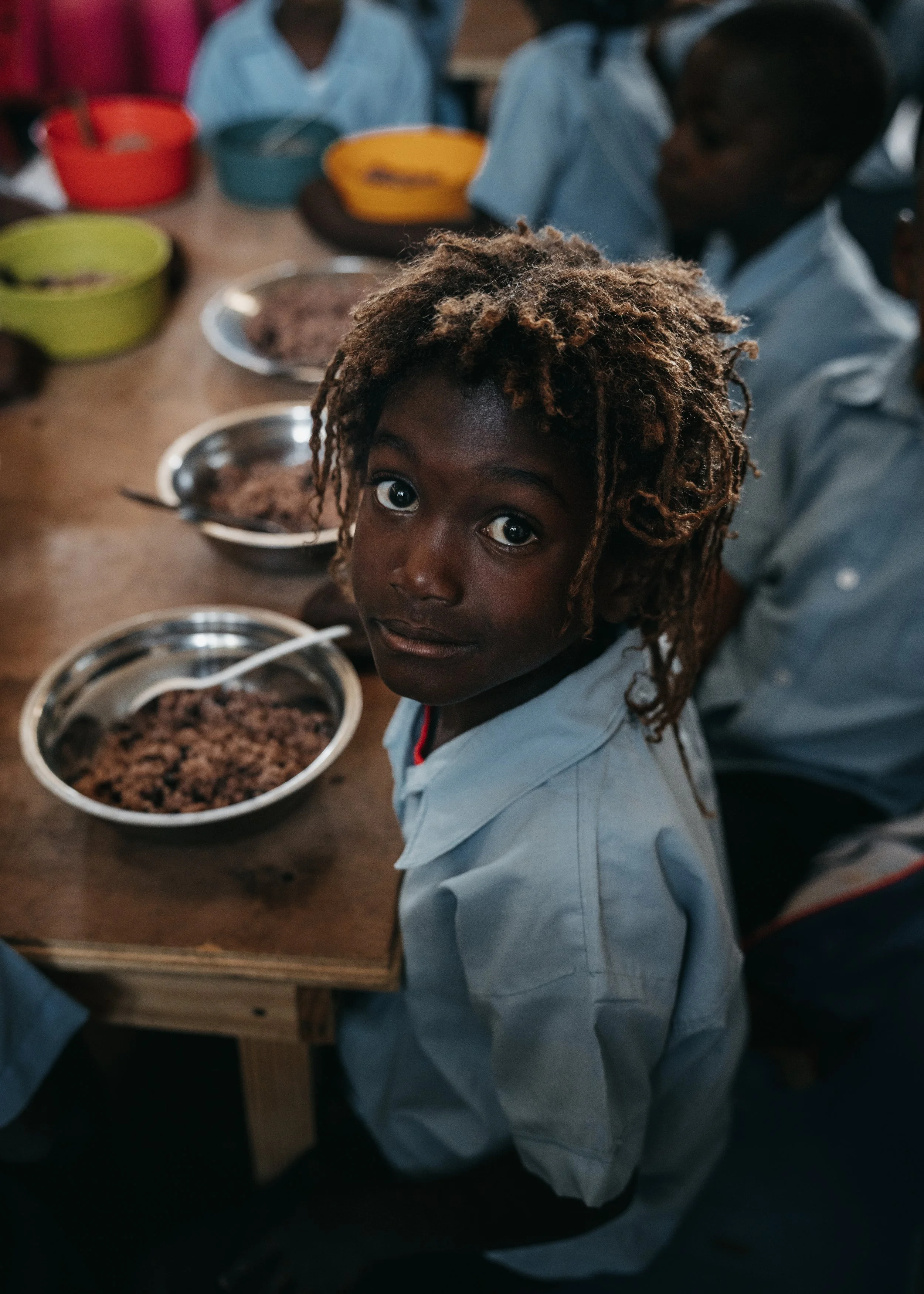 A young boy from Haiti with light brown, curly hair and dark skin looking at the camera while sitting at a wooden table with bowls of food, surrounded by other children in a classroom setting.