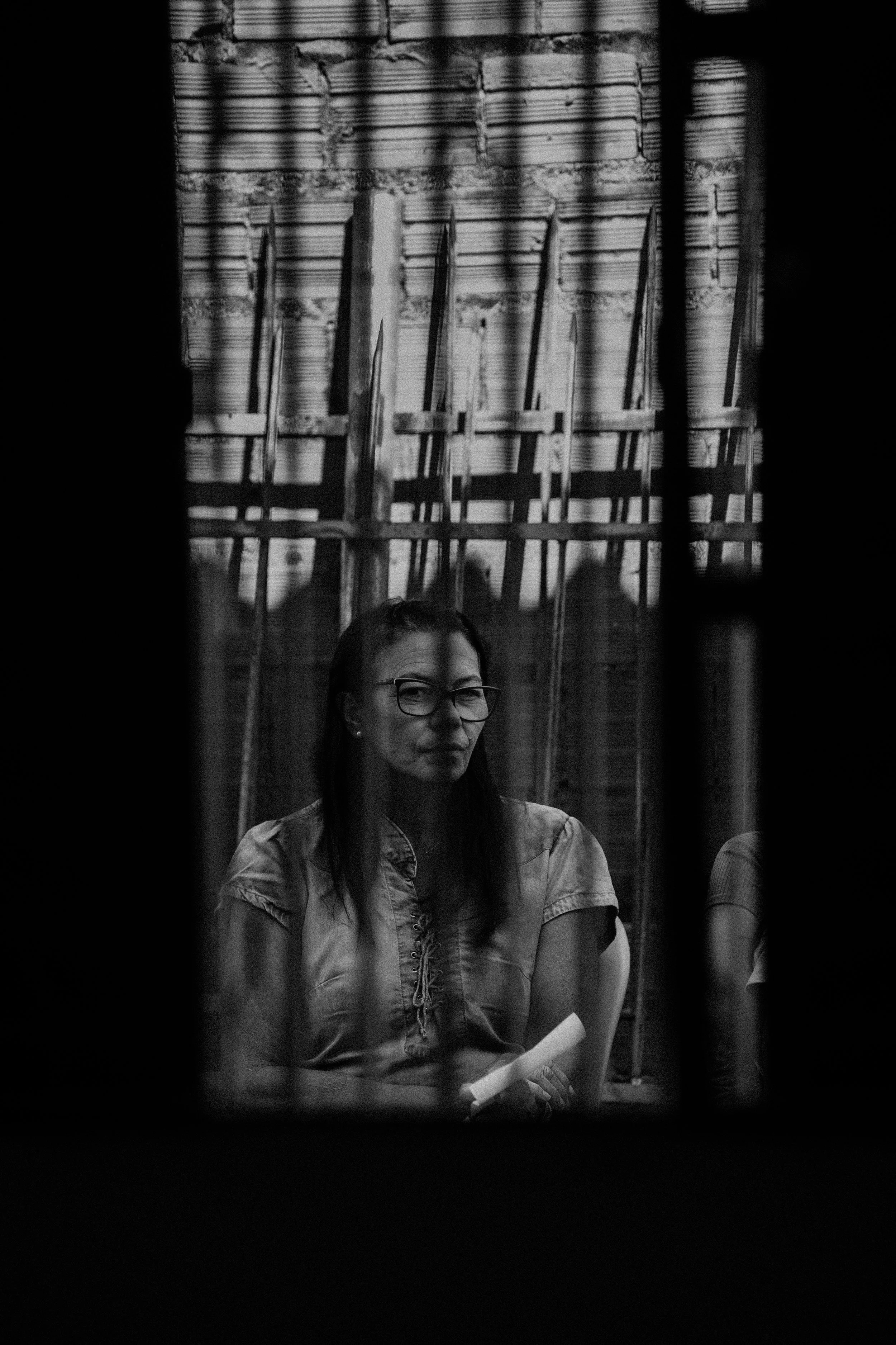 A woman with glasses and long dark hair sitting indoors, viewed through a window or opening with bars, in black and white.