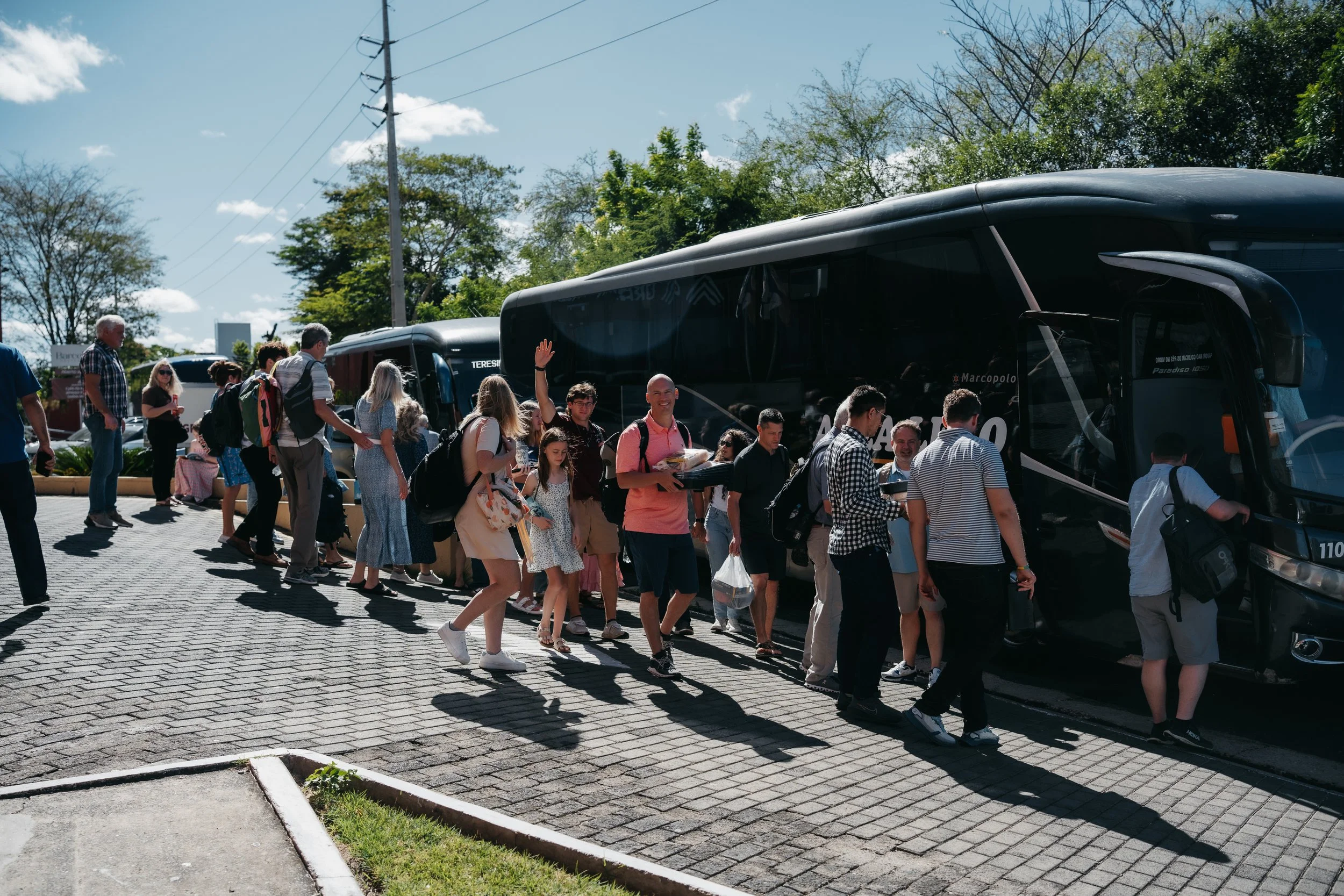 People boarding a black tour bus on a sunny day with green trees and blue sky in the background.