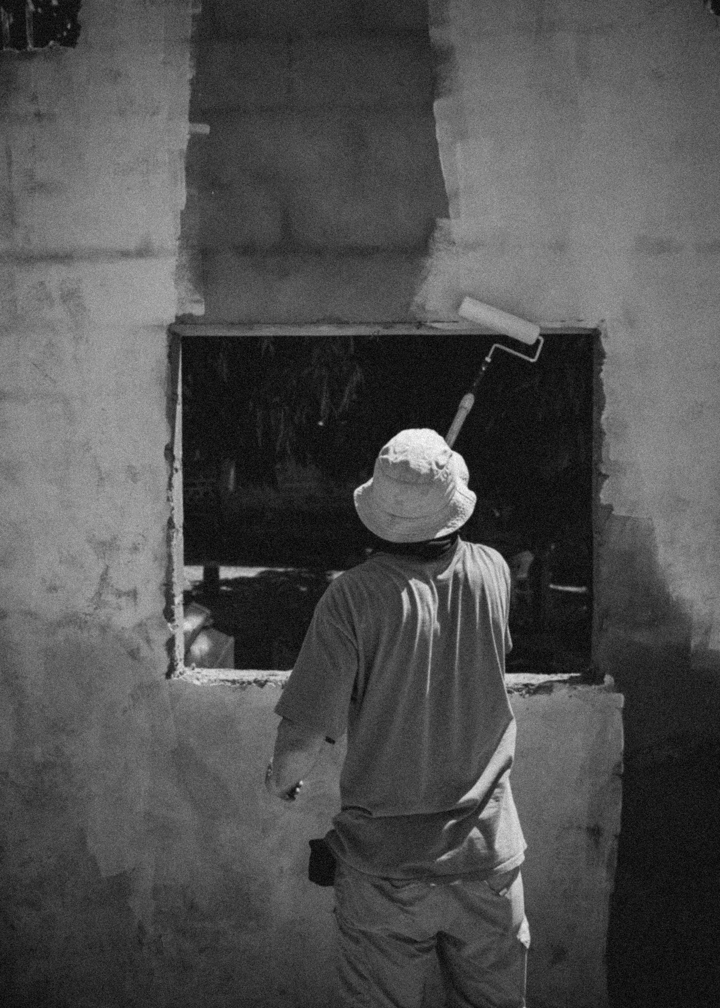Man wearing a bucket hat painting a window frame with a roller brush.