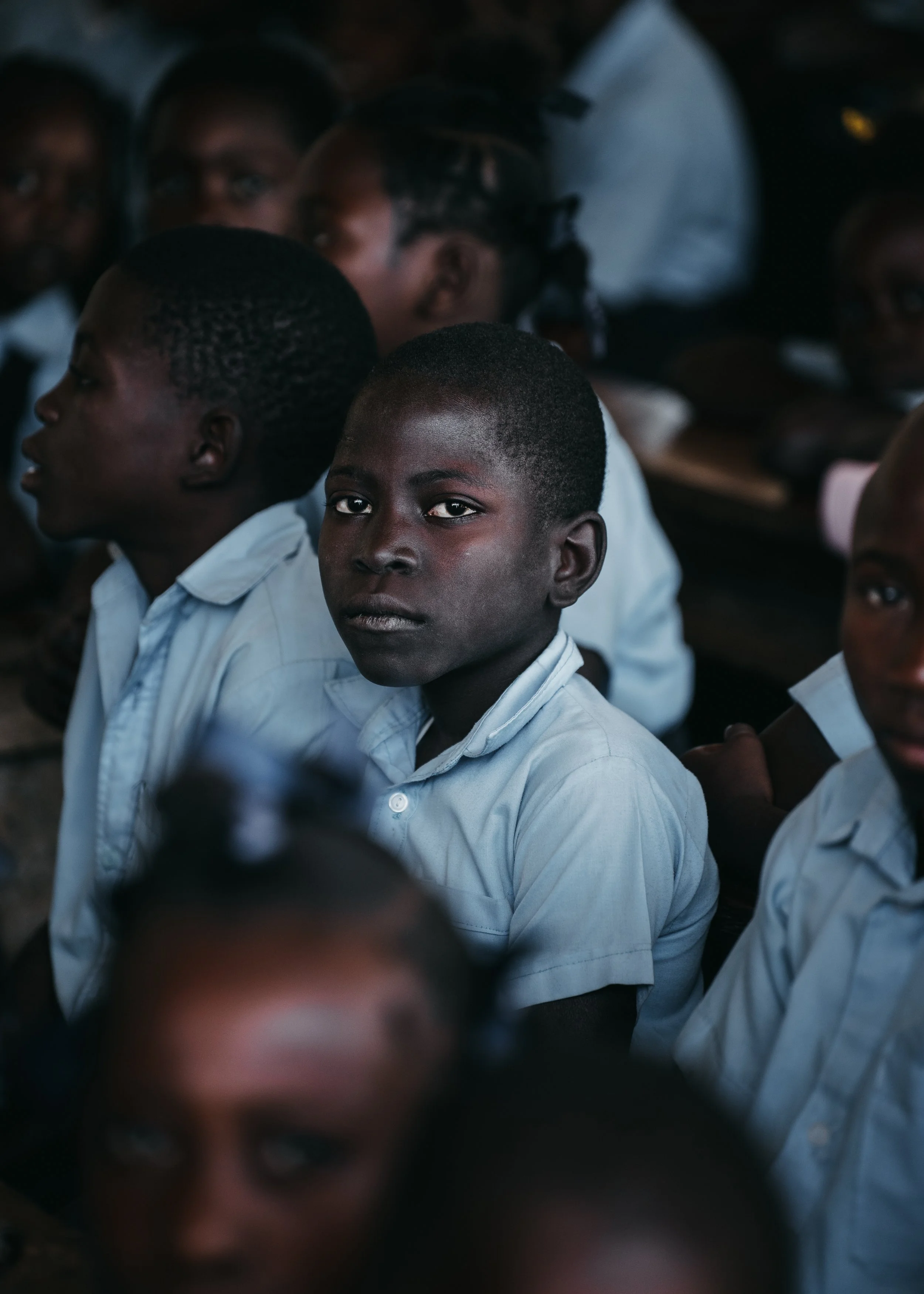 A group of young African students wearing white shirts, seated and looking towards the camera, with a focused and serious expression.