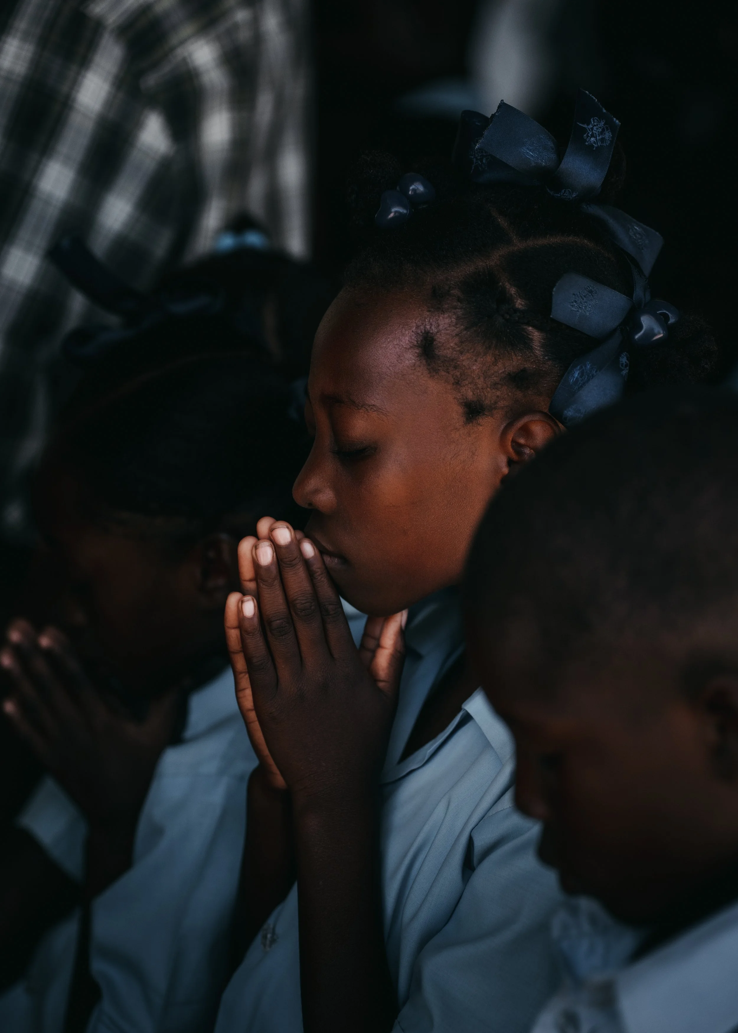 Close-up of a young girl with braided hair and blue bows, praying with her hands pressed together and eyes closed, surrounded by other children also praying.