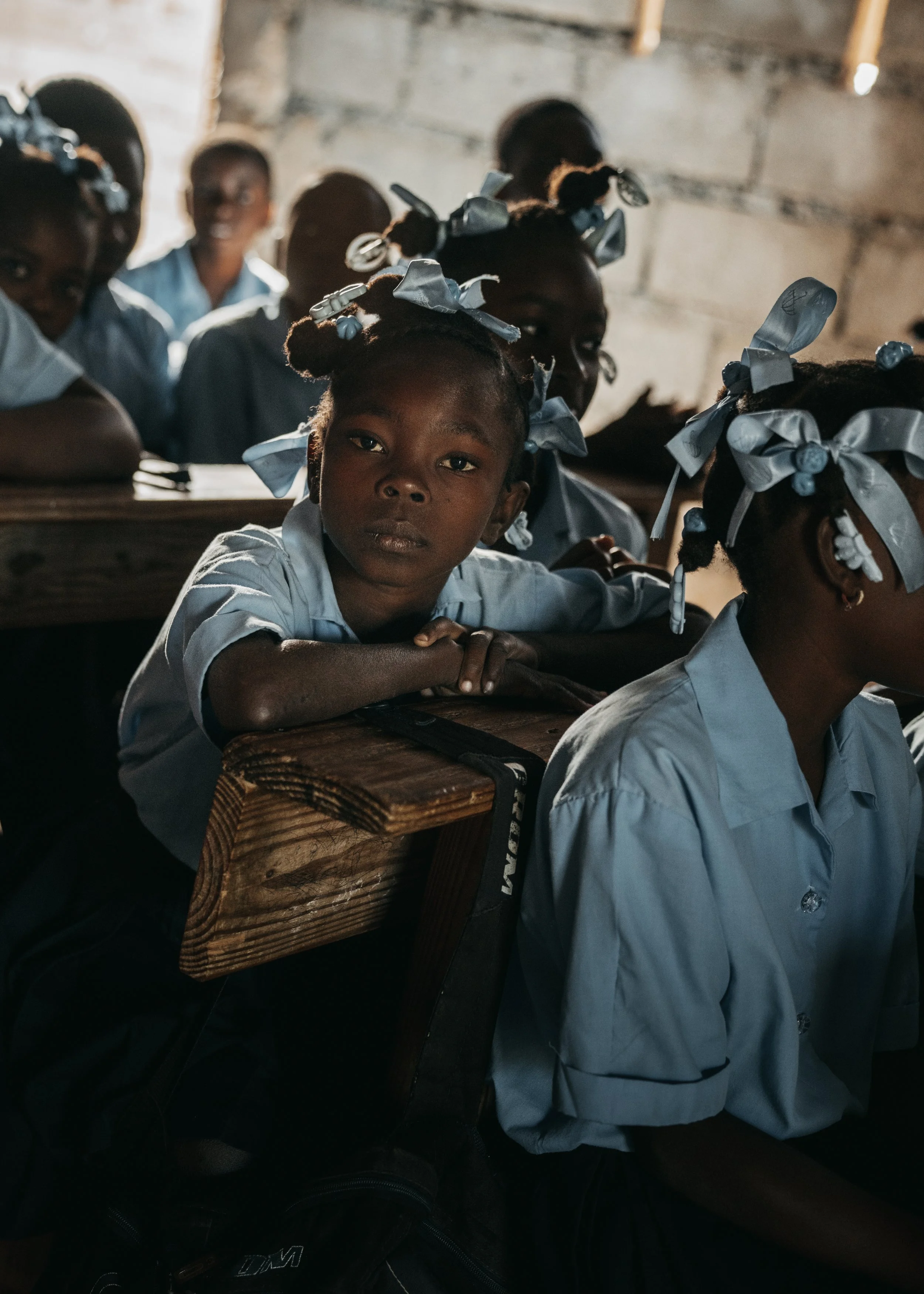 School children inside a classroom, some looking at the camera, wearing light blue uniforms and with blue ribbons in their hair, sitting at wooden desks.