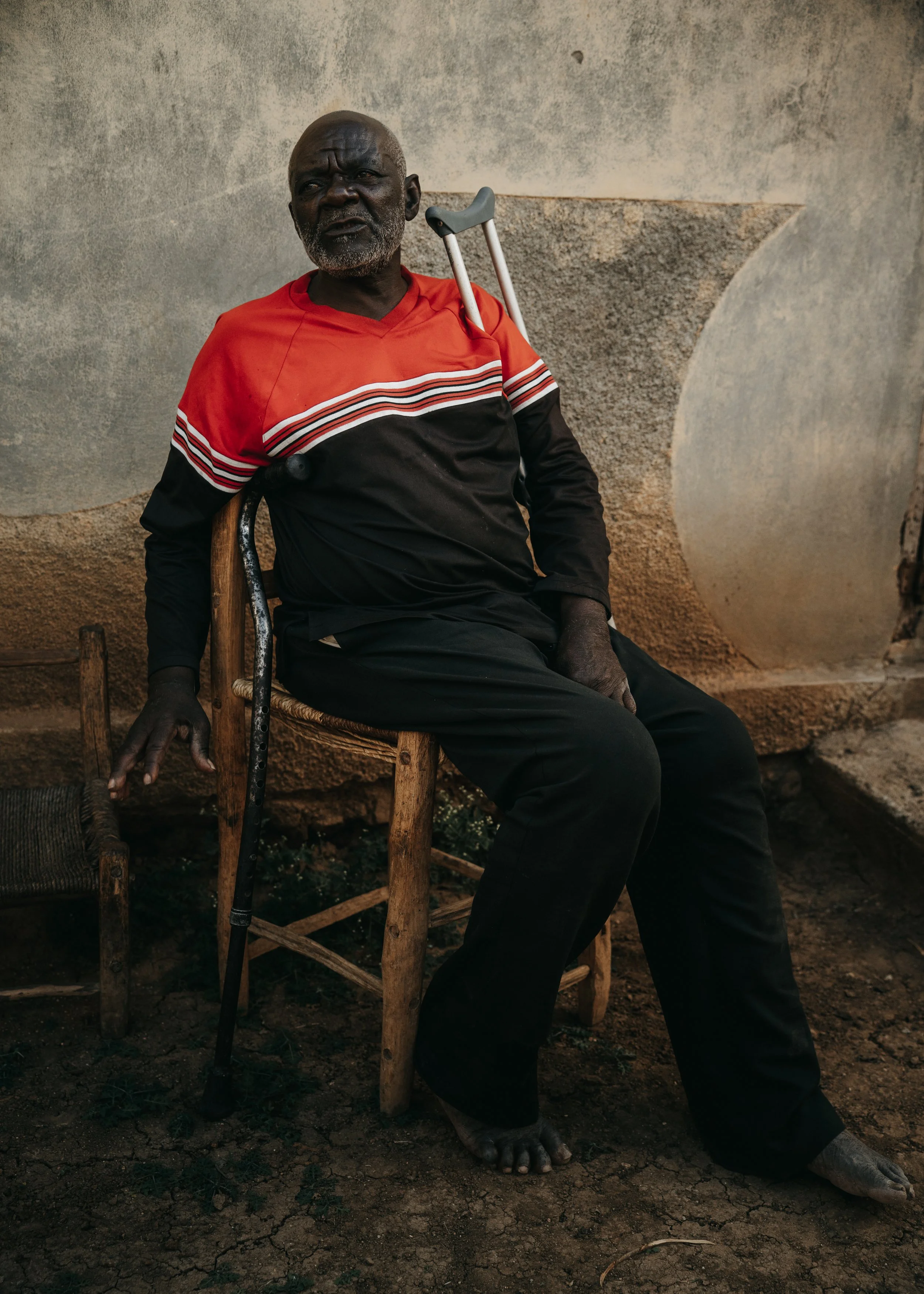 An elderly man with a beard sitting on a wooden chair against a textured wall, wearing a red and black shirt and dark pants, with a crutch resting on his shoulder and his foot resting on the ground.