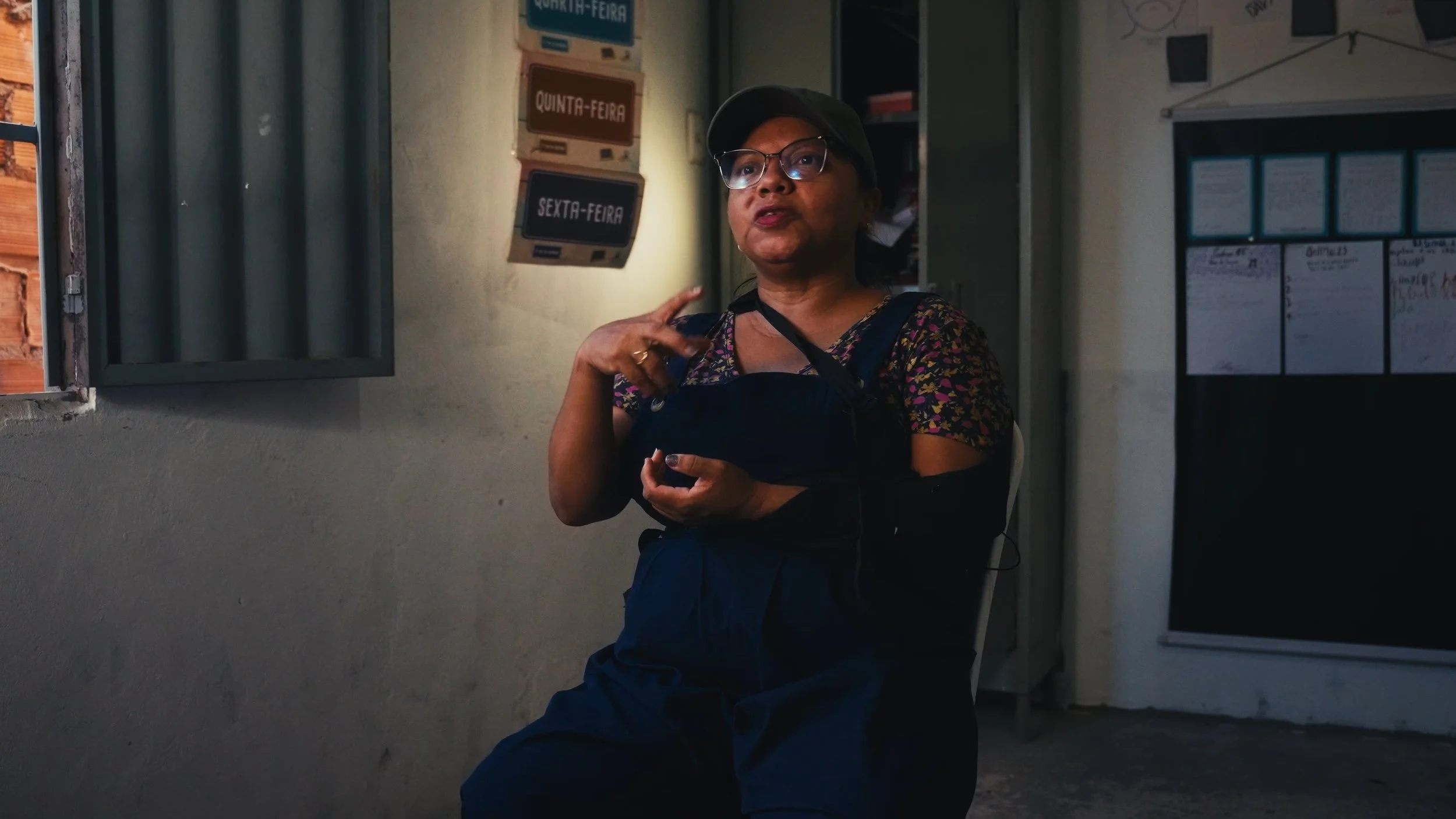 Woman with glasses and a hat sitting indoors near an open window, gesturing with her hand, with signs and a whiteboard on the wall behind her.