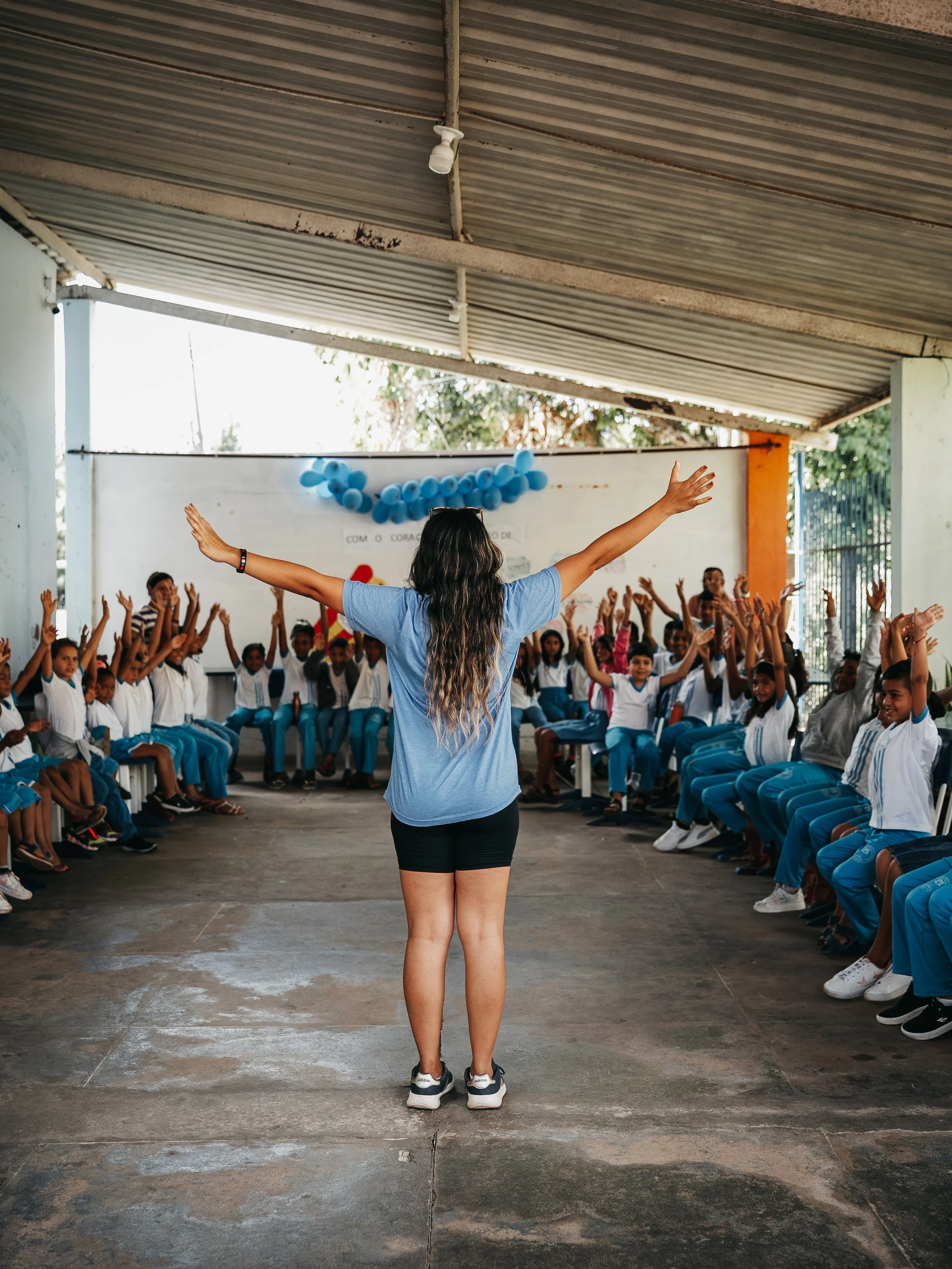 A woman with long brown hair wearing a blue t-shirt and black shorts leading a group of children in a seated activity under a covered outdoor area. The children are sitting in a semi-circle, raising their hands, and wearing school uniforms. There are blue balloons and a white banner with red and yellow lettering in the background.