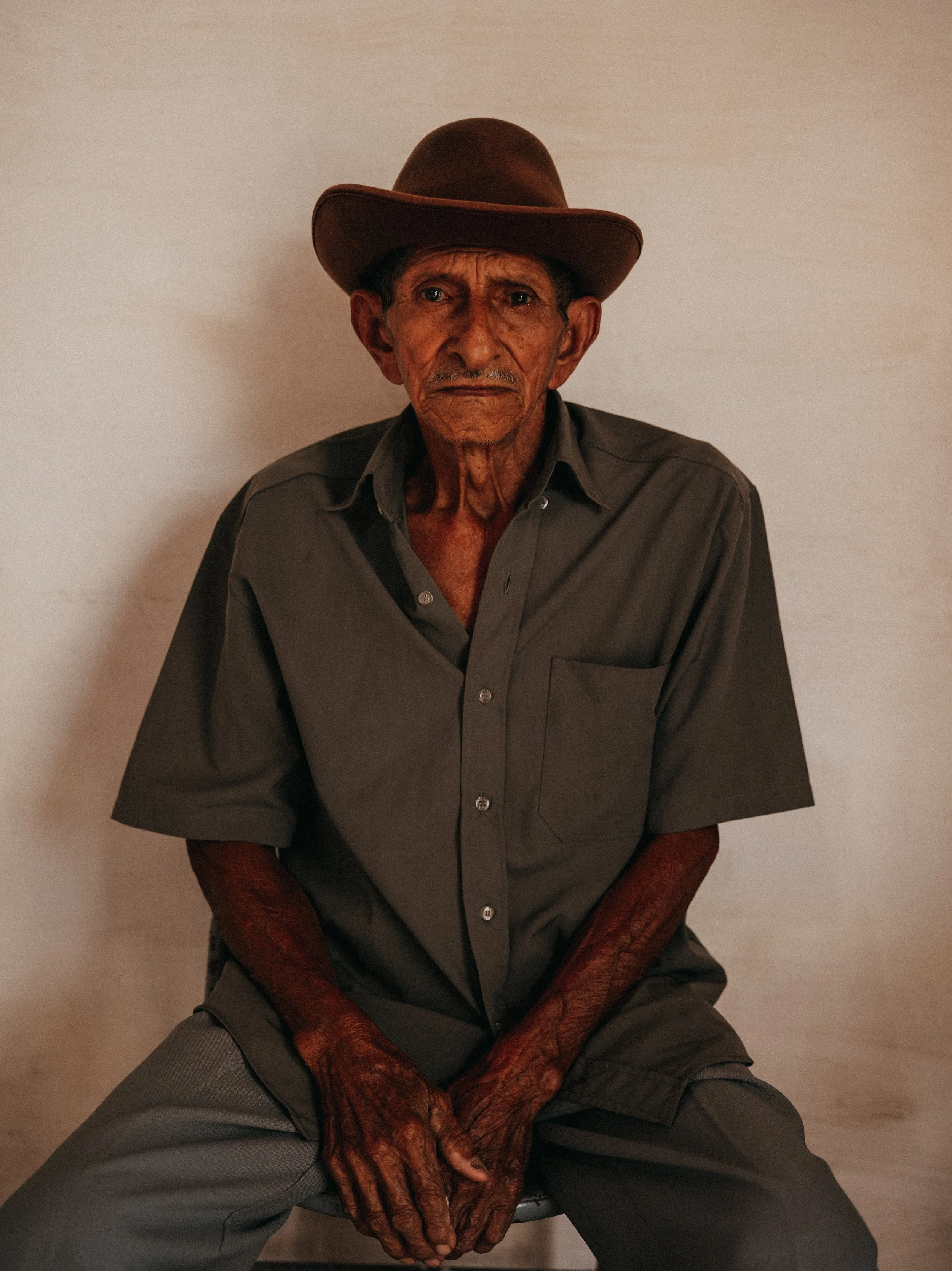An elderly man sitting against a plain light-colored wall, wearing a dark gray button-down shirt and a brown cowboy hat, with his hands resting on his lap and a serious expression on his face.