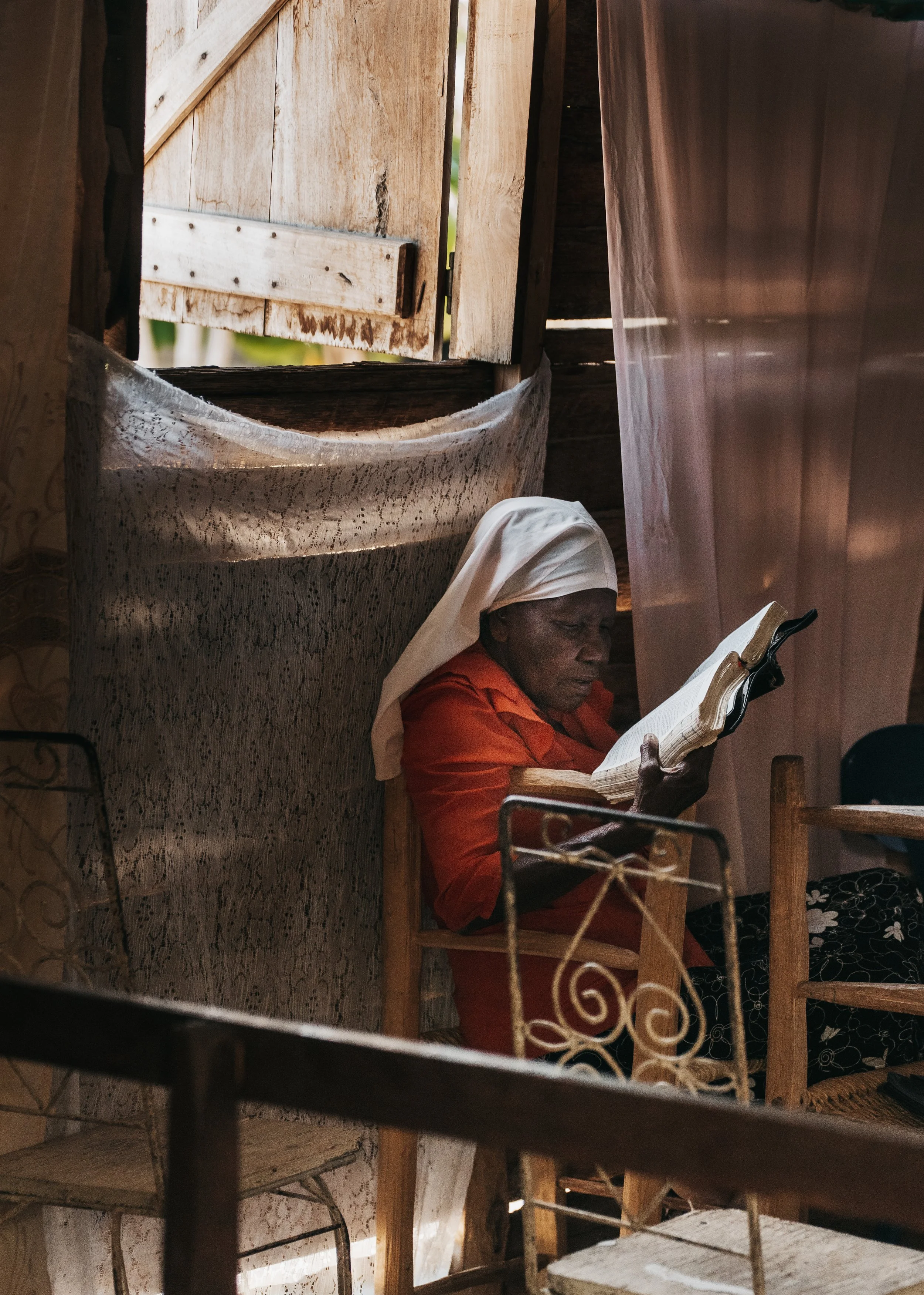 An elderly woman wearing a white headscarf and red blouse, sitting on a wooden chair inside a rustic wooden structure, reading a book.