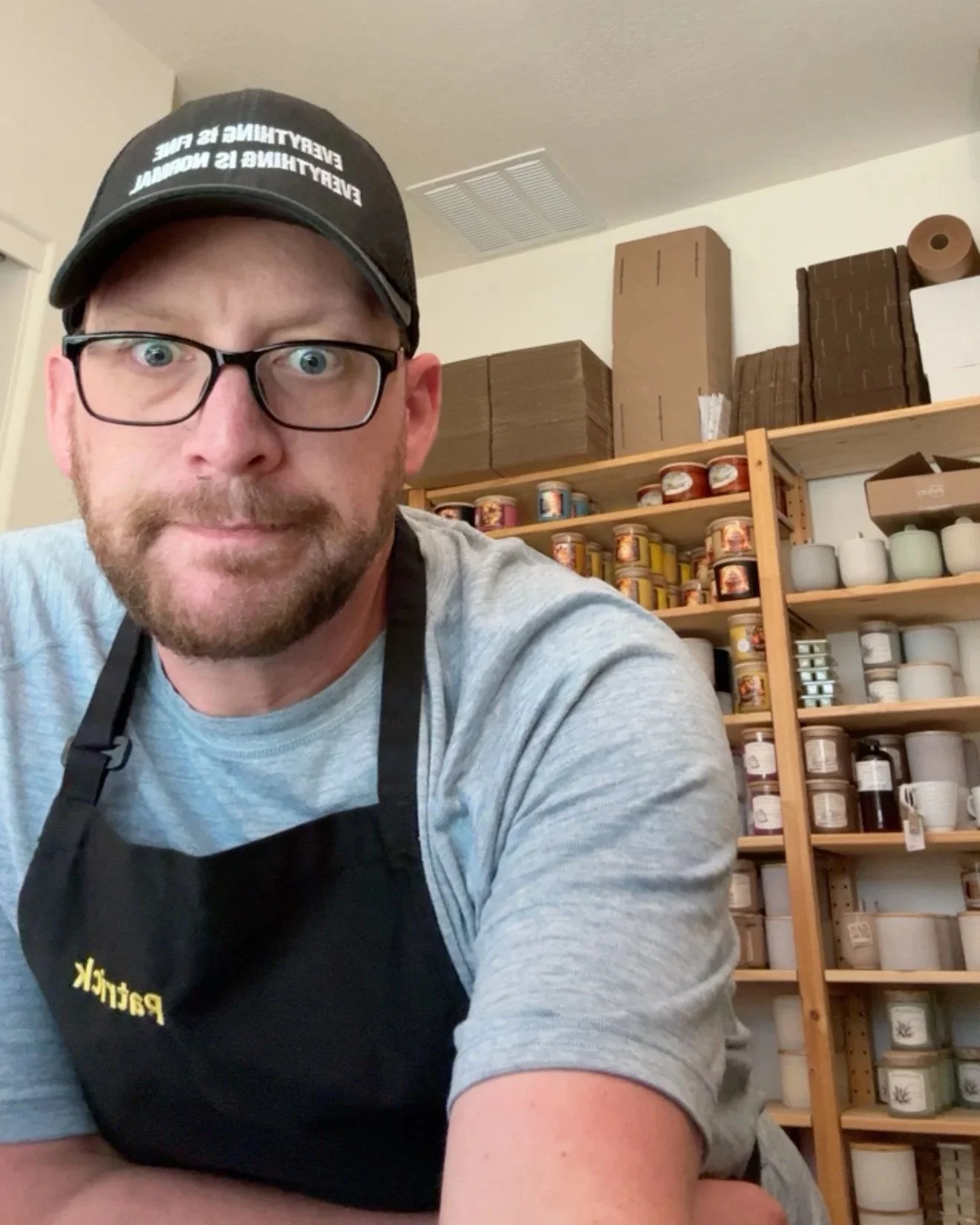 A man wearing glasses, a black cap, and a black apron, looking directly at the camera, with shelves of jars and boxes in the background, in what appears to be a storage room or pantry.