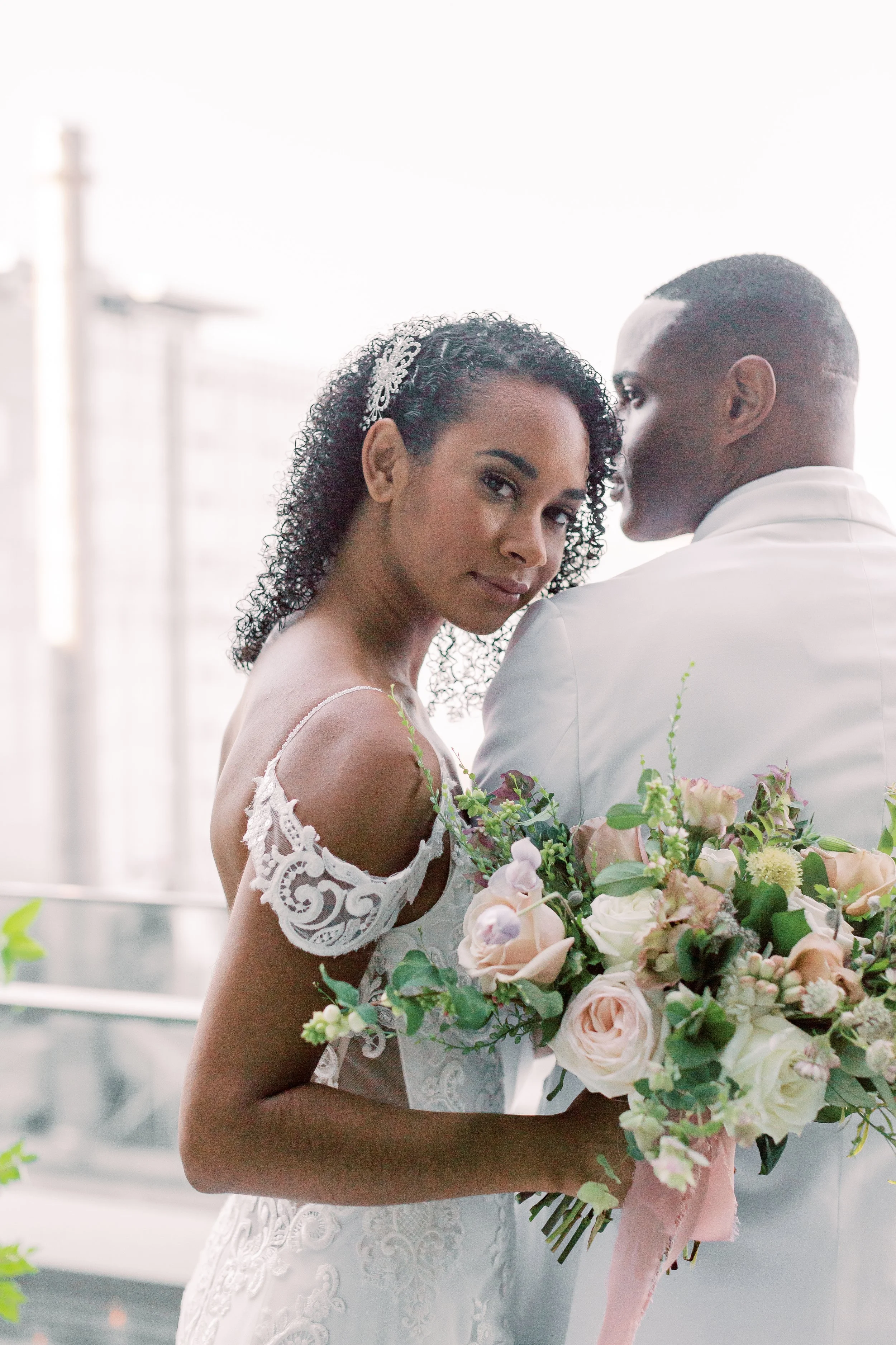 A bride and groom on their wedding day, with the bride holding a bouquet of pink and white flowers, standing close together and looking at the camera. The bride has curly hair and is wearing a lace dress, while the groom is dressed in a white suit.