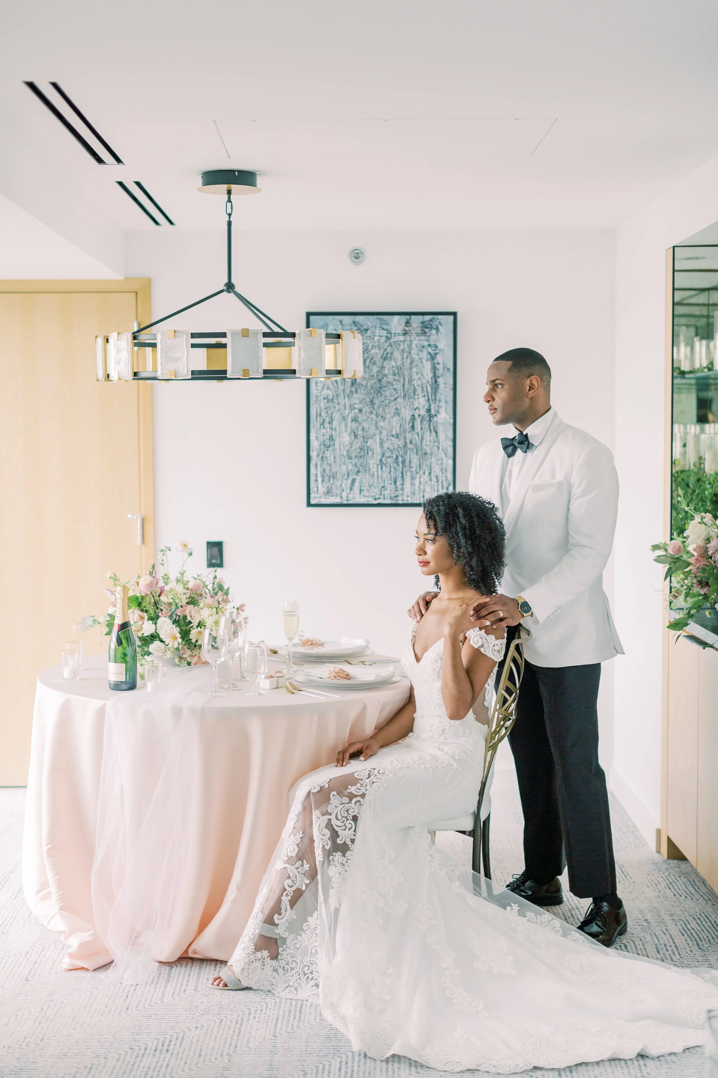 A bride in a white lace wedding dress seated at a decorated table with a groom in a white tuxedo standing behind her, indoors with modern decor.