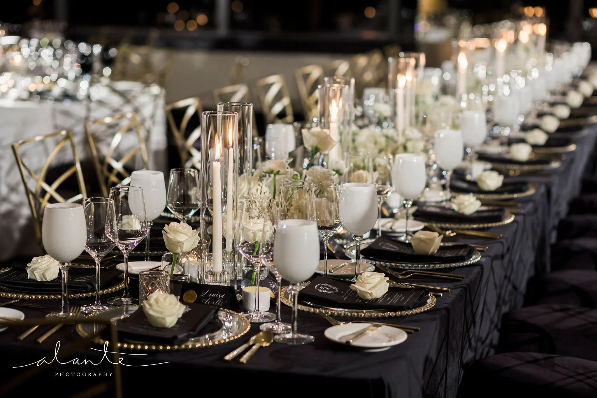 Elegant banquet table decorated with white roses, candles, black napkins, and gold flatware, set for a formal event.