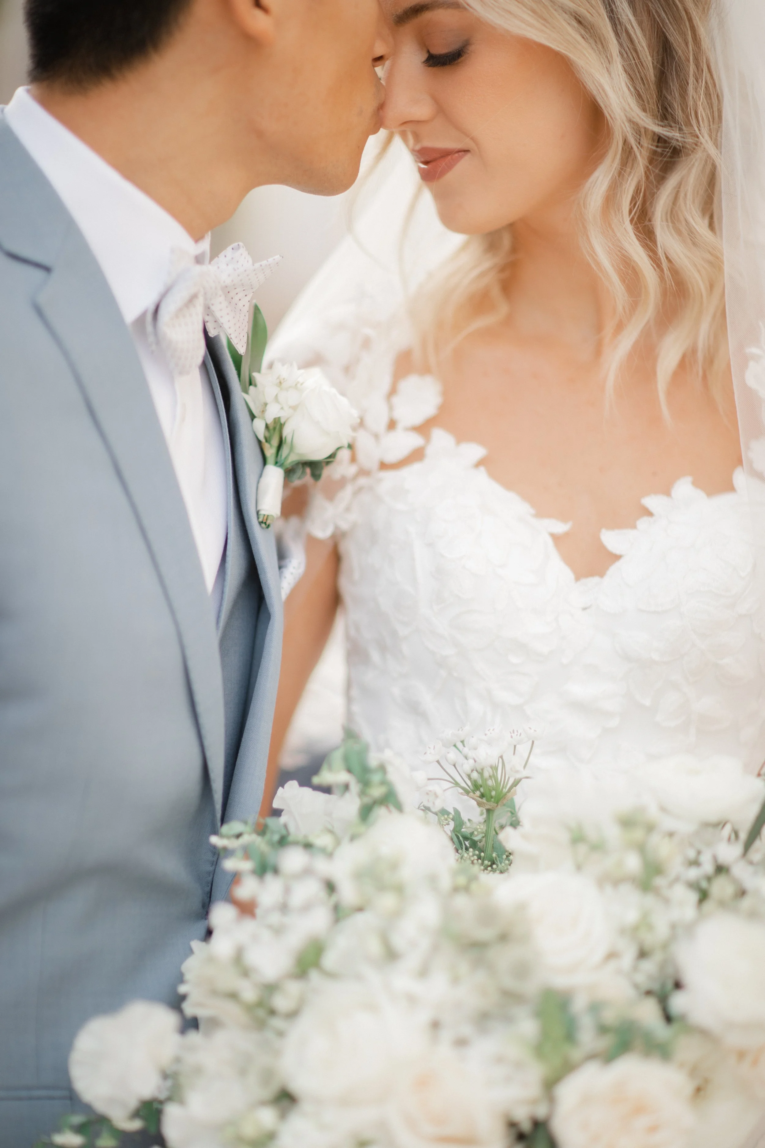 Close-up of a bride and groom touching foreheads with eyes closed, standing next to each other surrounded by wedding flowers.