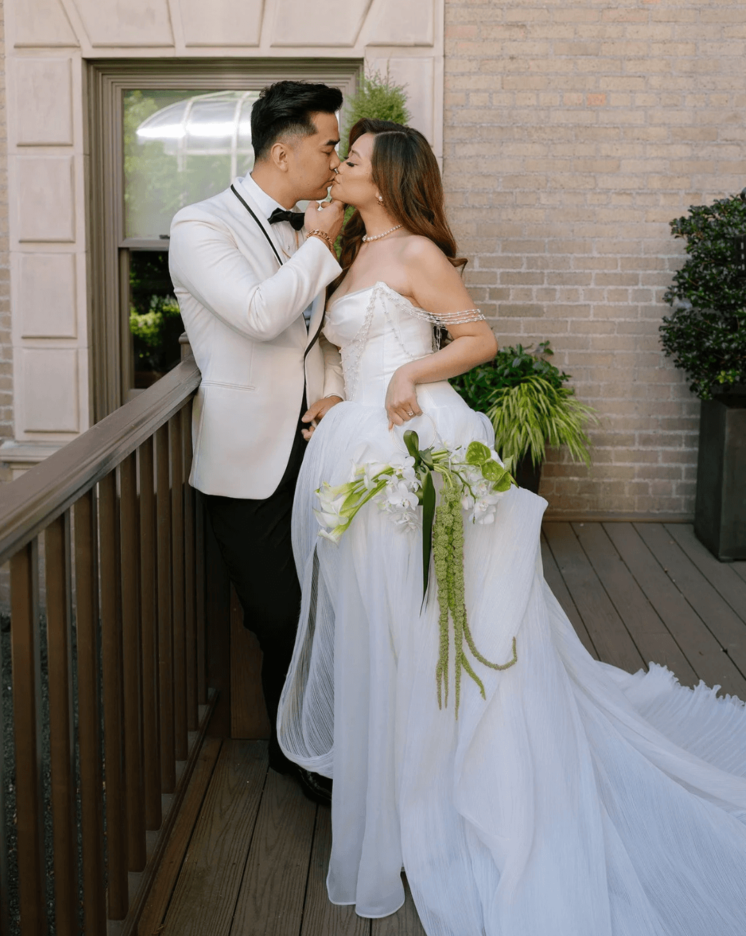 Bride and groom kissing on a balcony in formal attire with cascading bouquet at wedding venue