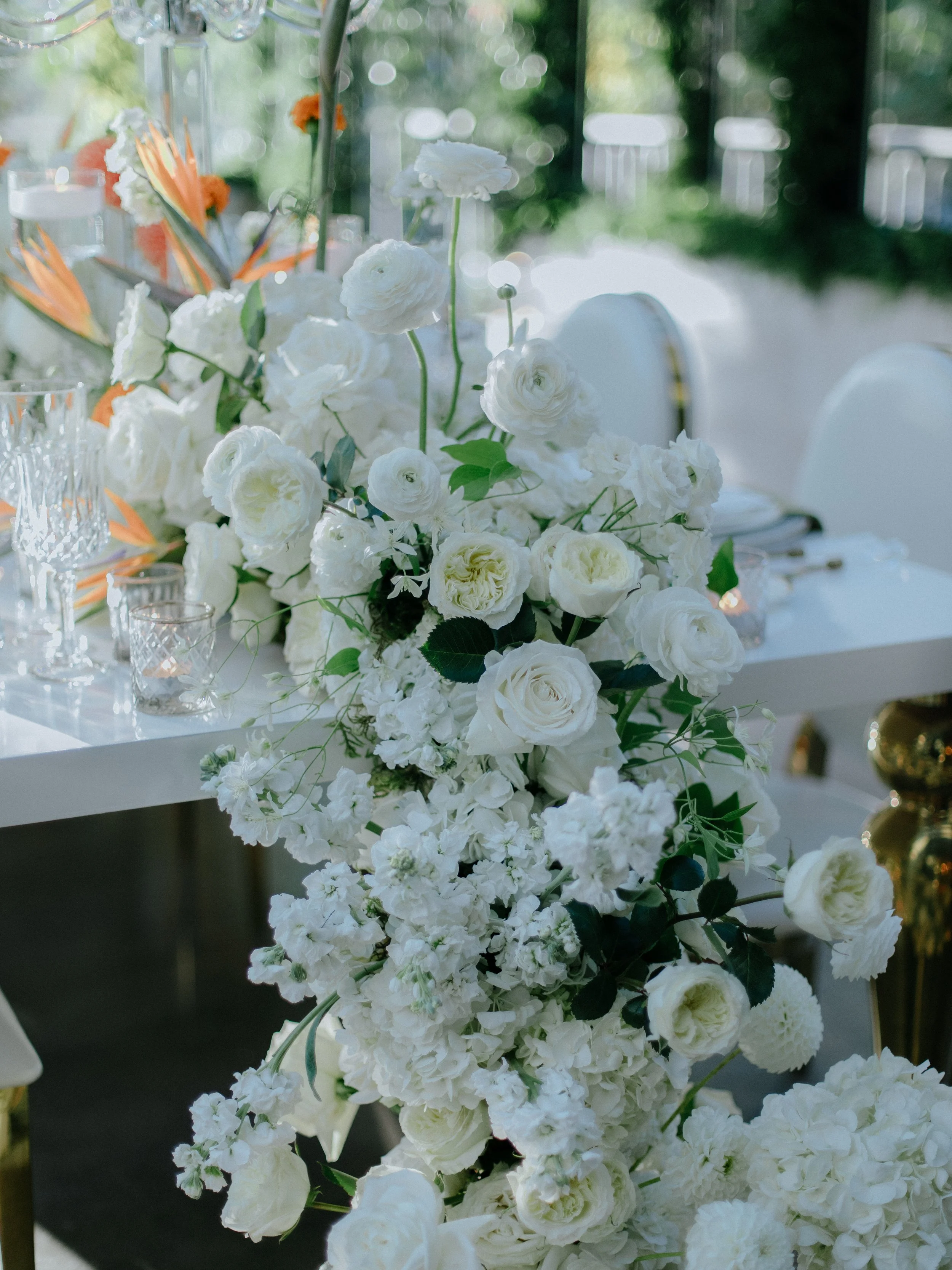 White floral centerpiece with roses, ranunculus, and greenery on a table at an outdoor event.