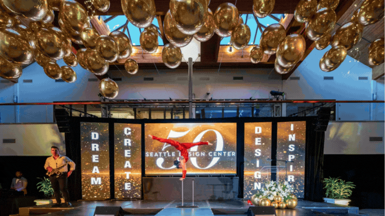 A performer in a red outfit is doing a handstand on a pole on stage at the Seattle Design Center's 50th anniversary celebration, with gold balloons hanging from the ceiling and decorative plants on stage.