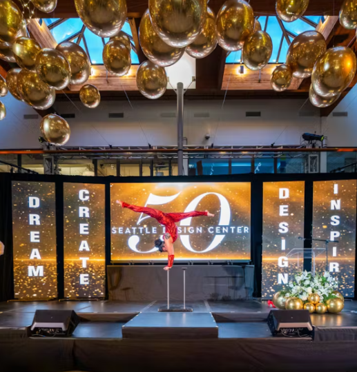 A performer doing an acrobatic pole dance on stage at the Seattle Design Center, decorated with gold ornaments and banners reading 'Dream Big,' 'Create,' 'Design,' 'Inspire,' and celebrating a 50th anniversary.