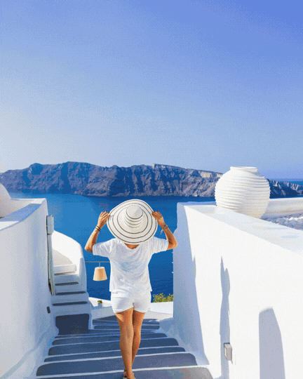 A woman in a white dress holding a large sunhat, walking down stairs on a balcony overlooking blue water and a distant island in Santorini, Greece.