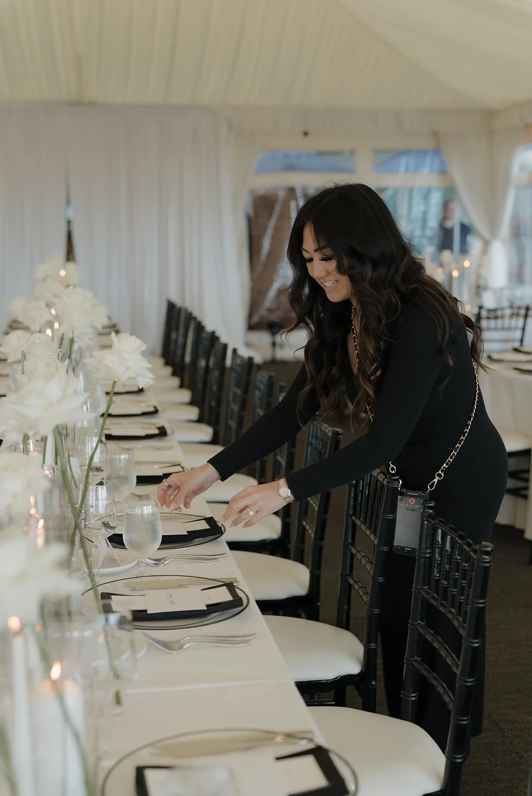 Woman in black dress setting a dining table with white flowers, glasses, and black napkins in a decorated event space.