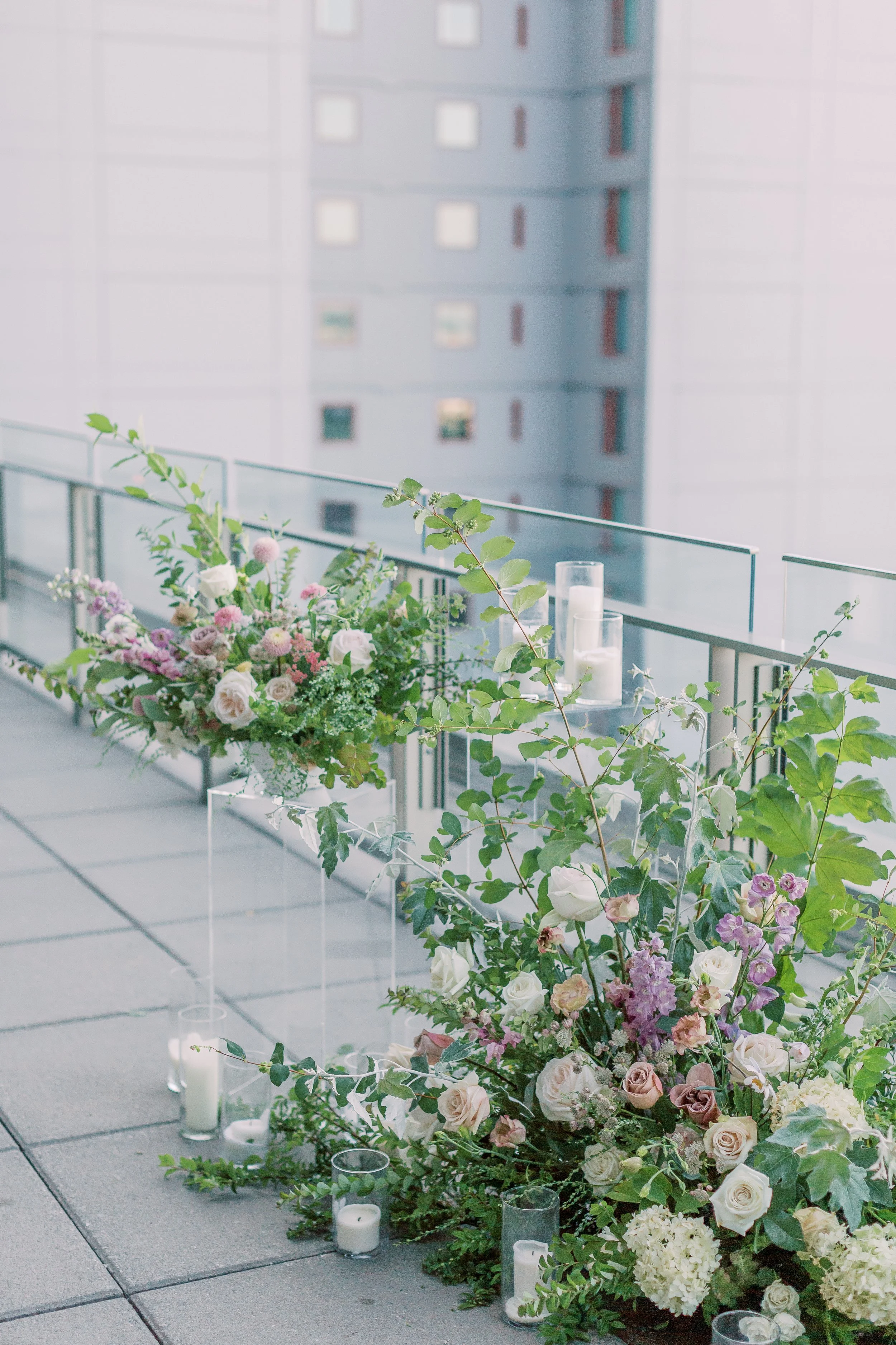 Floral arrangement with roses, hydrangeas, and greenery on a balcony with candles, overlooking a city building.