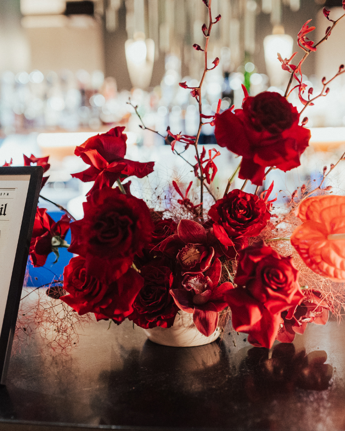 A bouquet of red roses and orchids in a white vase on a dark tabletop, with blurred background of a well-lit indoor space.