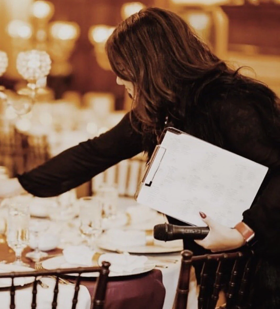 A woman setting a formal dining table with glassware and tableware, holding a clipboard and a microphone, in an elegant, warmly lit event space.