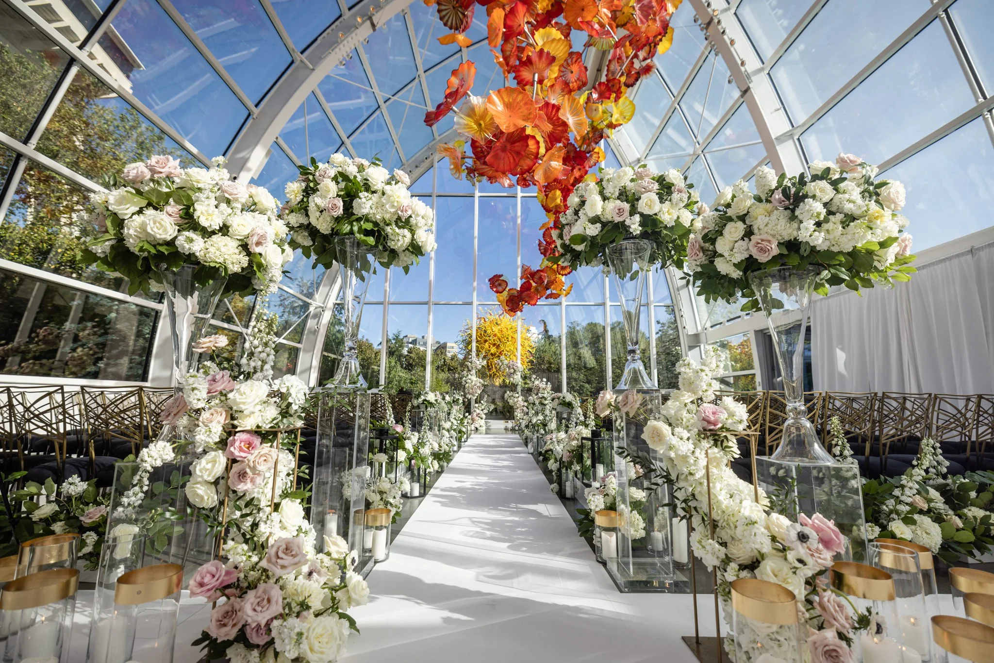 A wedding aisle inside a glass dome, decorated with tall floral arrangements of white and pink roses, placed in clear vases on tables, and an orange and yellow floral installation hanging from the ceiling.