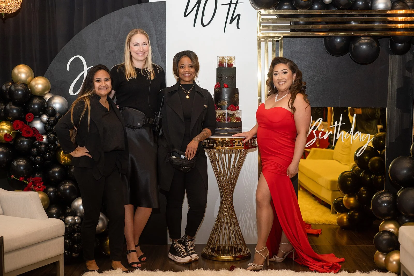 Four women standing in front of a decorated wall with the words '40th Birthday.' One woman in a red dress with a high slit stands next to a table holding a tiered cake decorated with black, red, and gold accents. The women are smiling, and the background features black, gold, and silver balloons and a yellow couch in a nearby room.