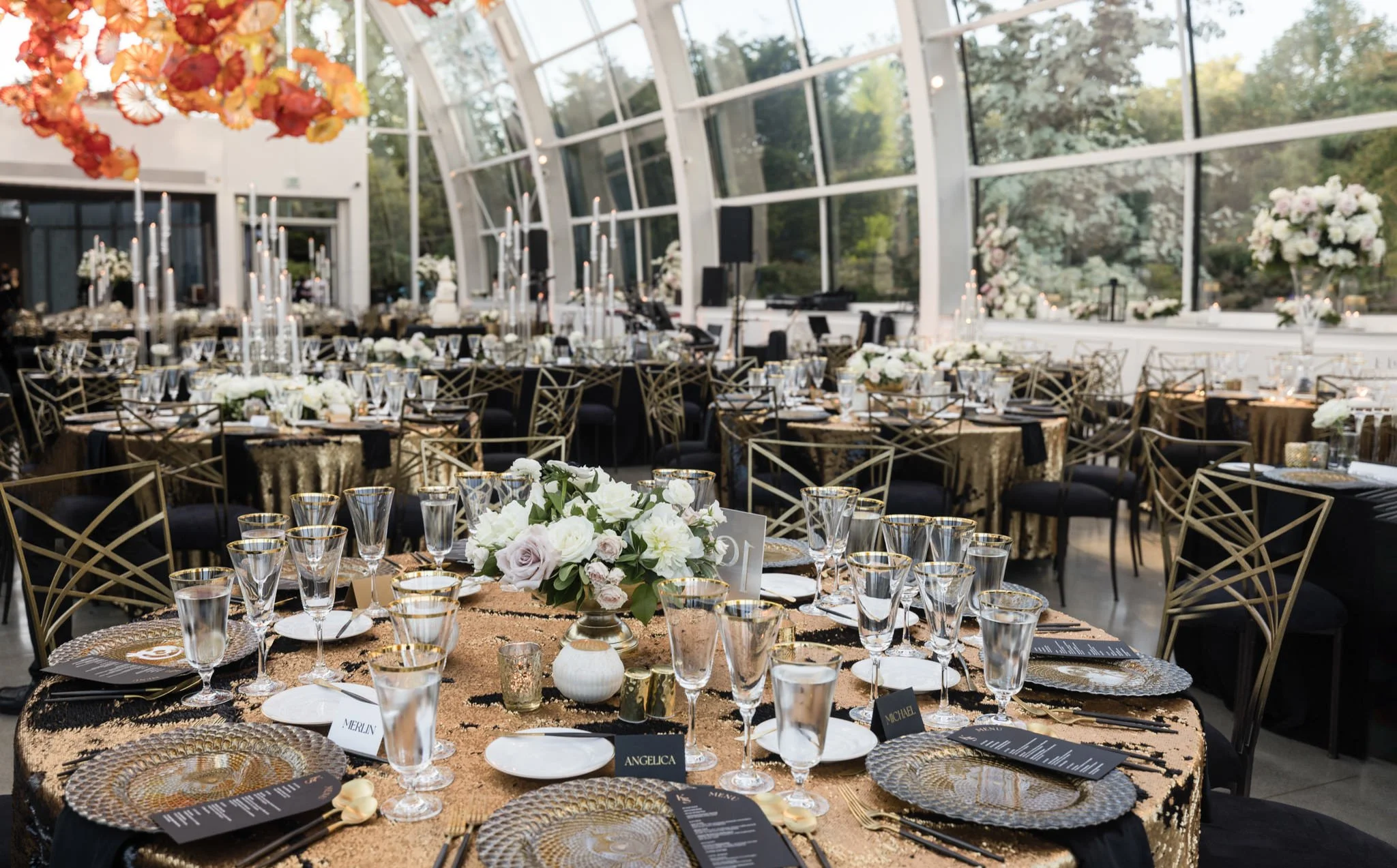 Elegant indoor wedding reception setup with round tables, gold and black chairs, and floral centerpieces, in a glass-walled venue with a high arched ceiling and natural light.