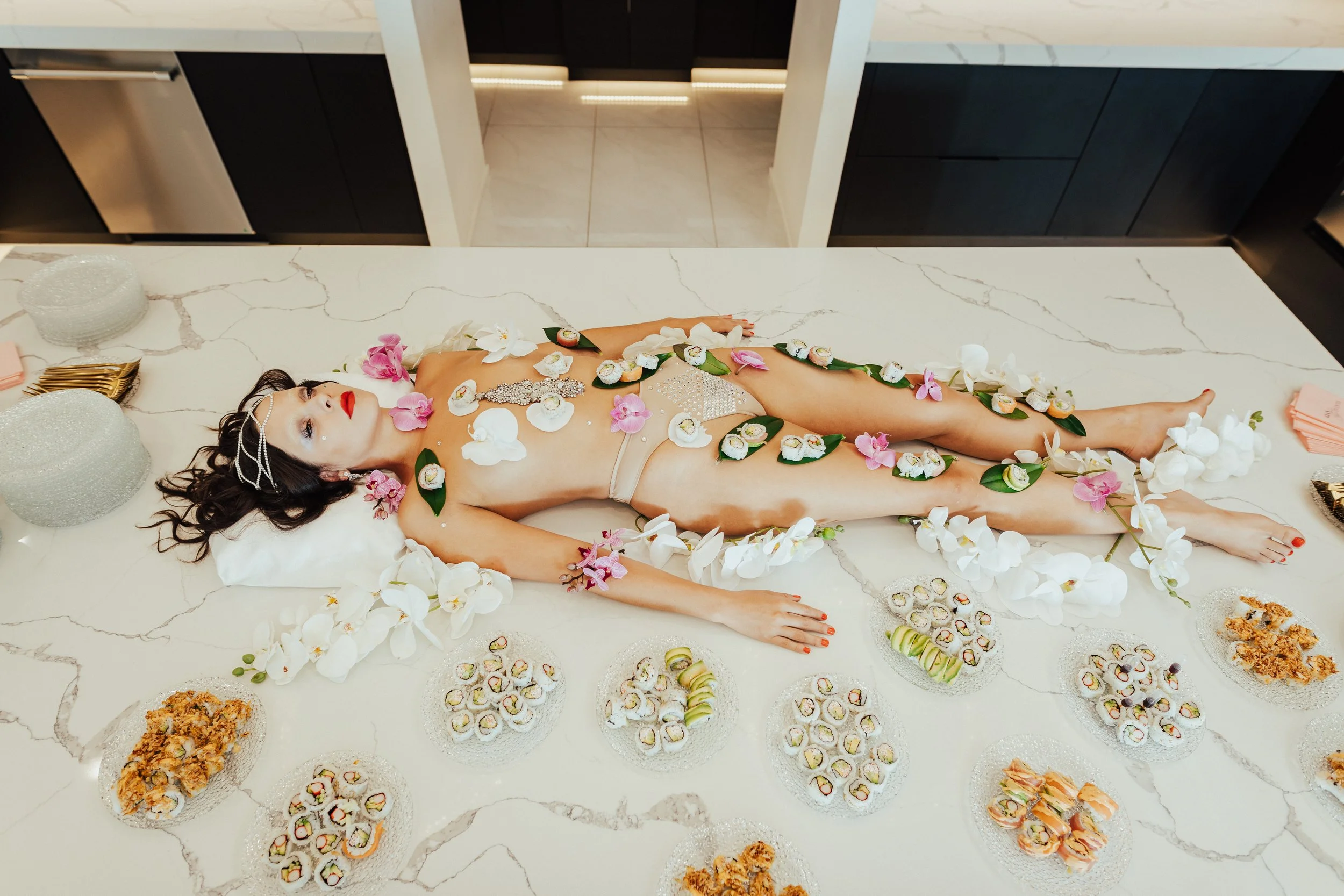 A woman lying naked on a marble kitchen island covered with white and pink orchids, sushi, and other appetizers, surrounded by a modern kitchen setting.