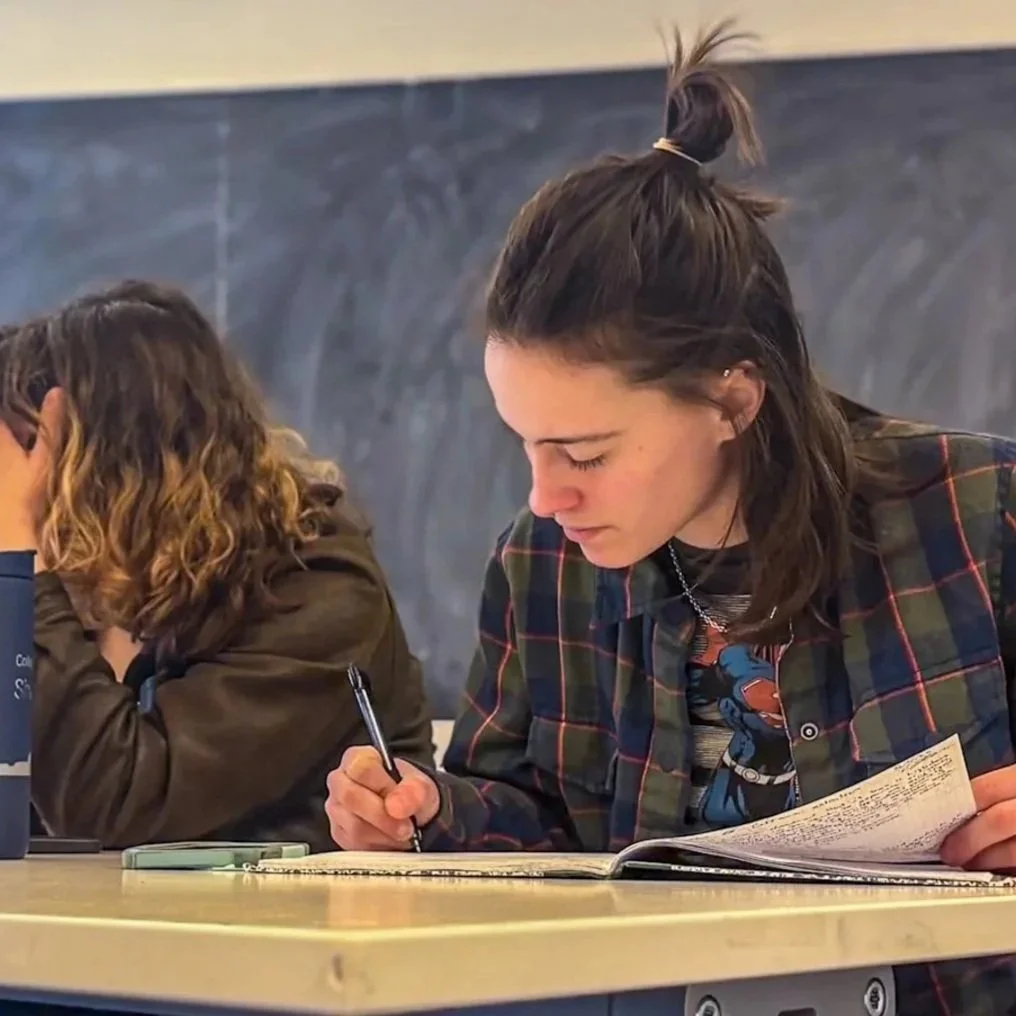 students writing in notebooks at a table. A chalkboard is in the background.