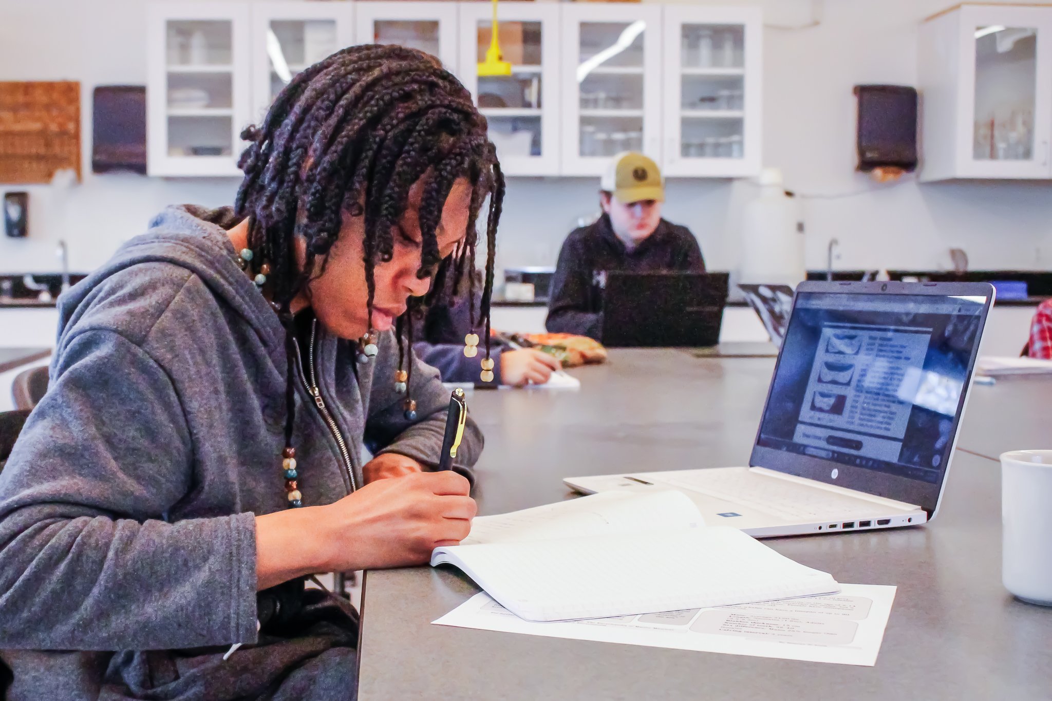 Students are writing in notebooks and reading on laptops at a table in a classroom lab.
