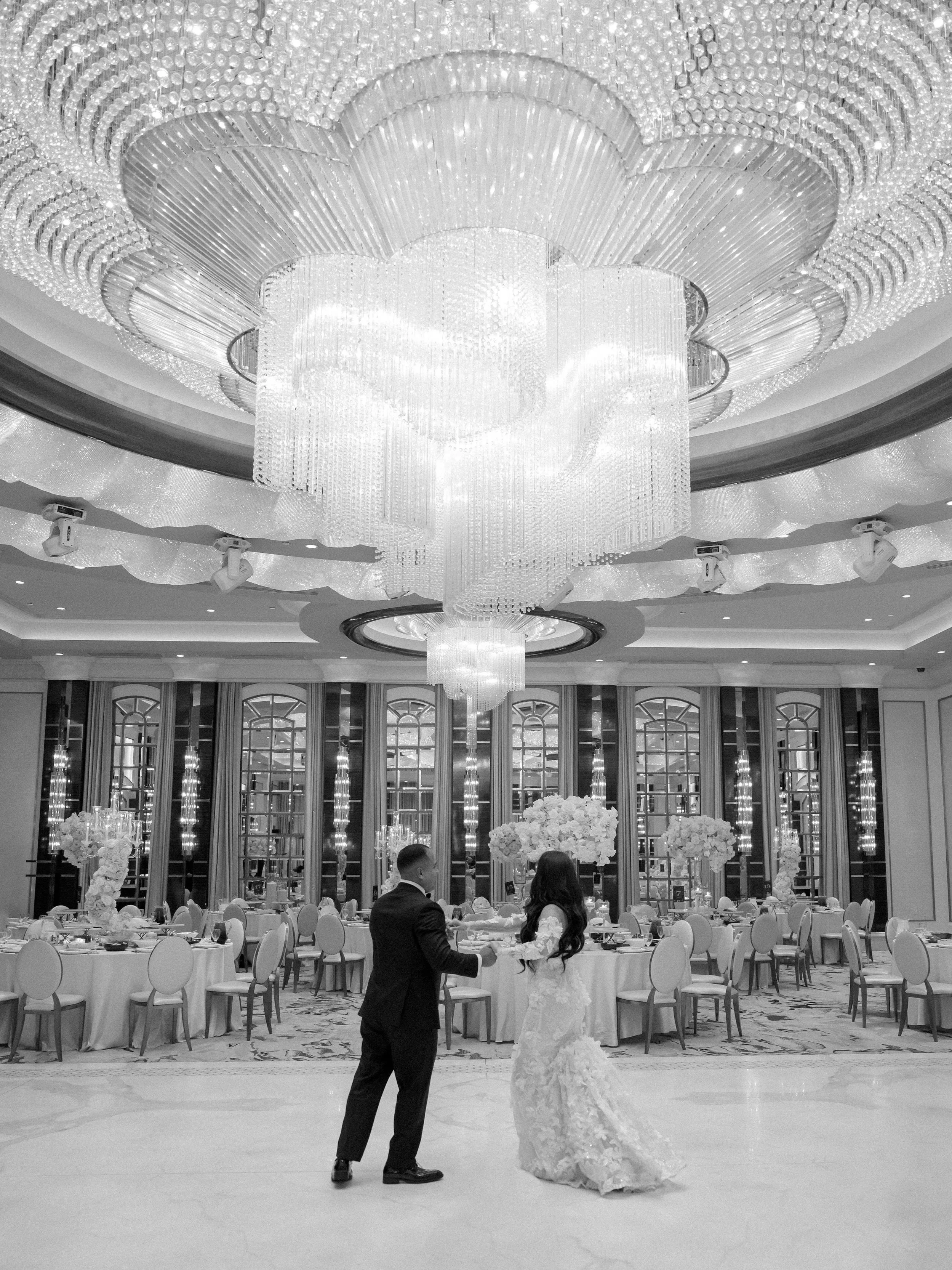 A bride and groom dance in a luxurious wedding reception hall with a large, ornate chandelier hanging from the ceiling. The hall features round tables with floral centerpieces and elegant decor.