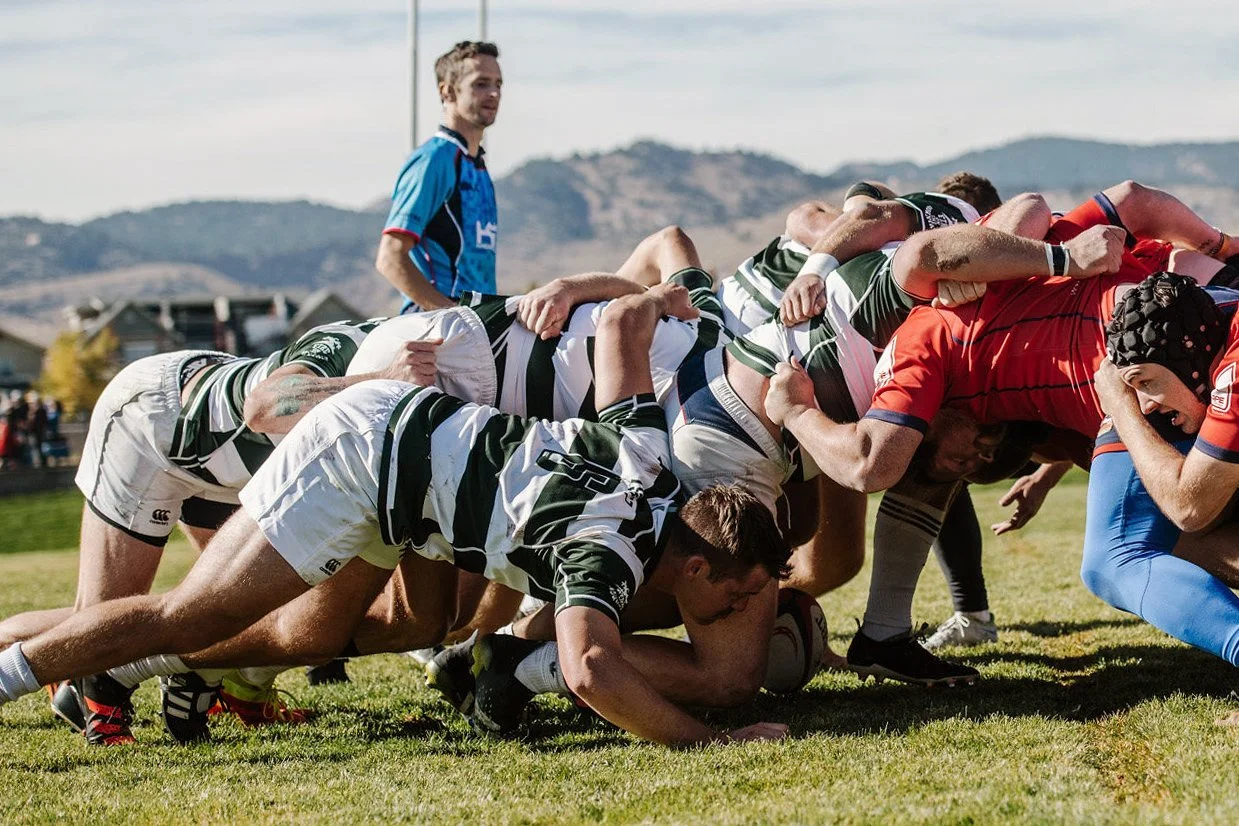 Rugby players in a scrum during a match, with a referee observing in the background.