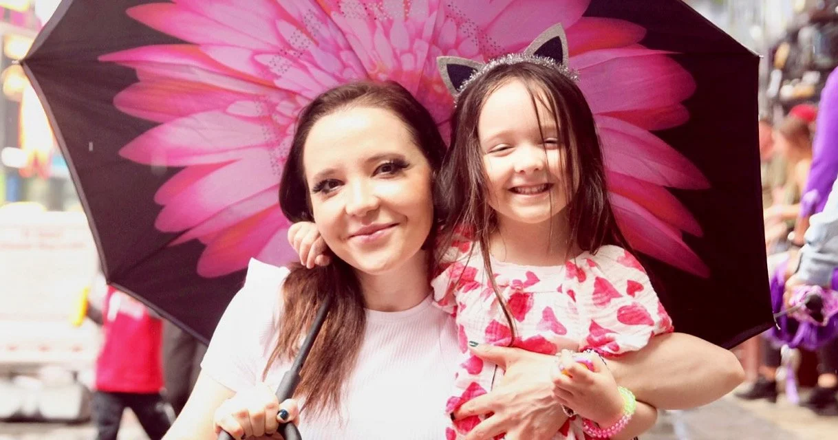 A woman and a young girl smiling under a pink flower-patterned umbrella in a crowded outdoor setting.