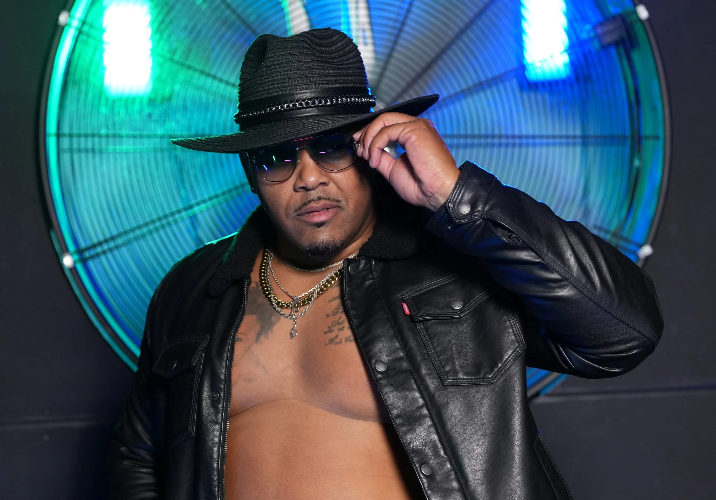 A man wearing sunglasses, a black leather jacket, and a large black hat posing in front of a colorful illuminated circular background.