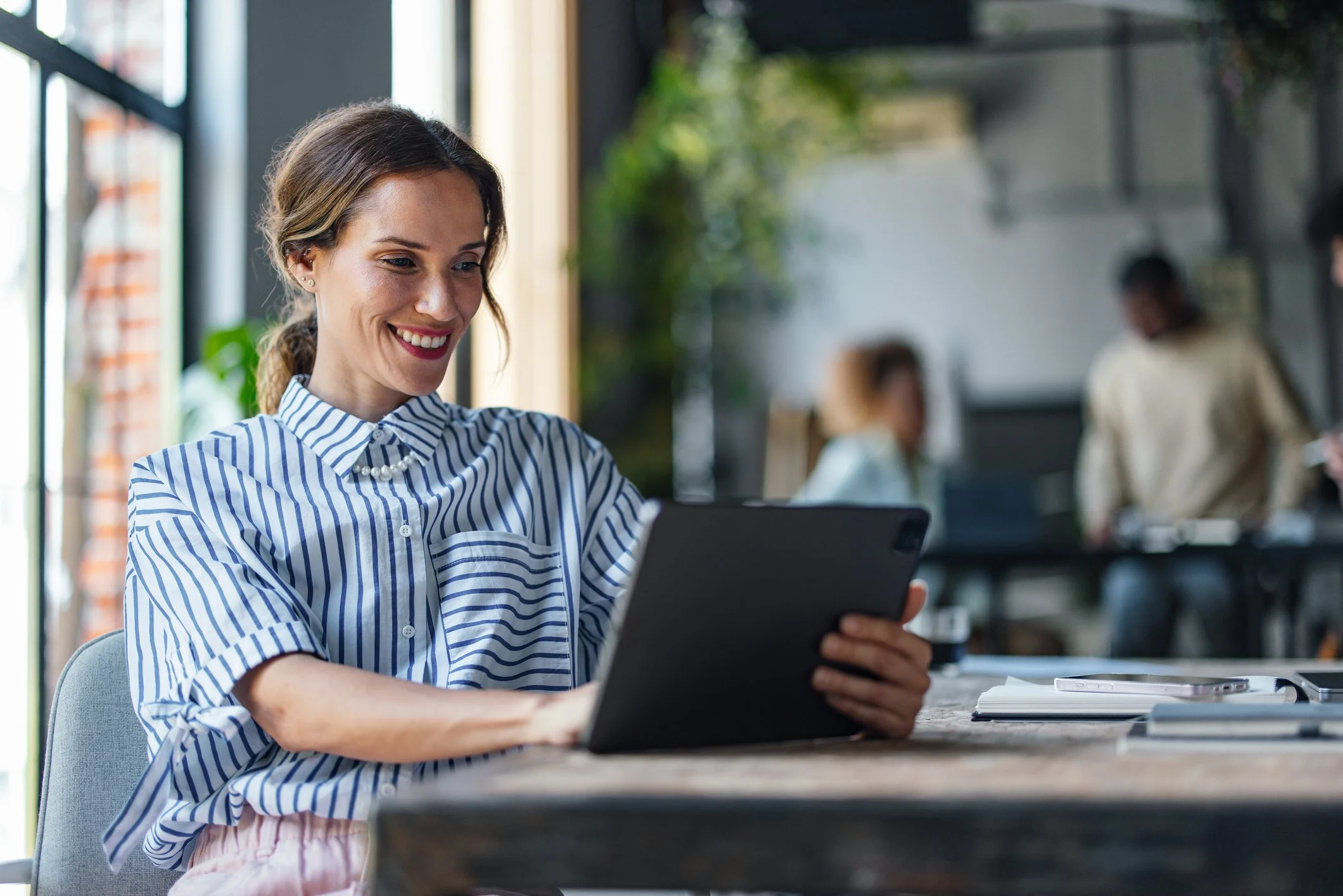 A young woman with brown hair tied back, wearing a blue and white striped shirt and a pearl necklace, sits at a table using a tablet. She is smiling and appears to be engaging with the device. In the background, there are other people working and socializing in a modern, well-lit indoor space with large windows and green plants.
