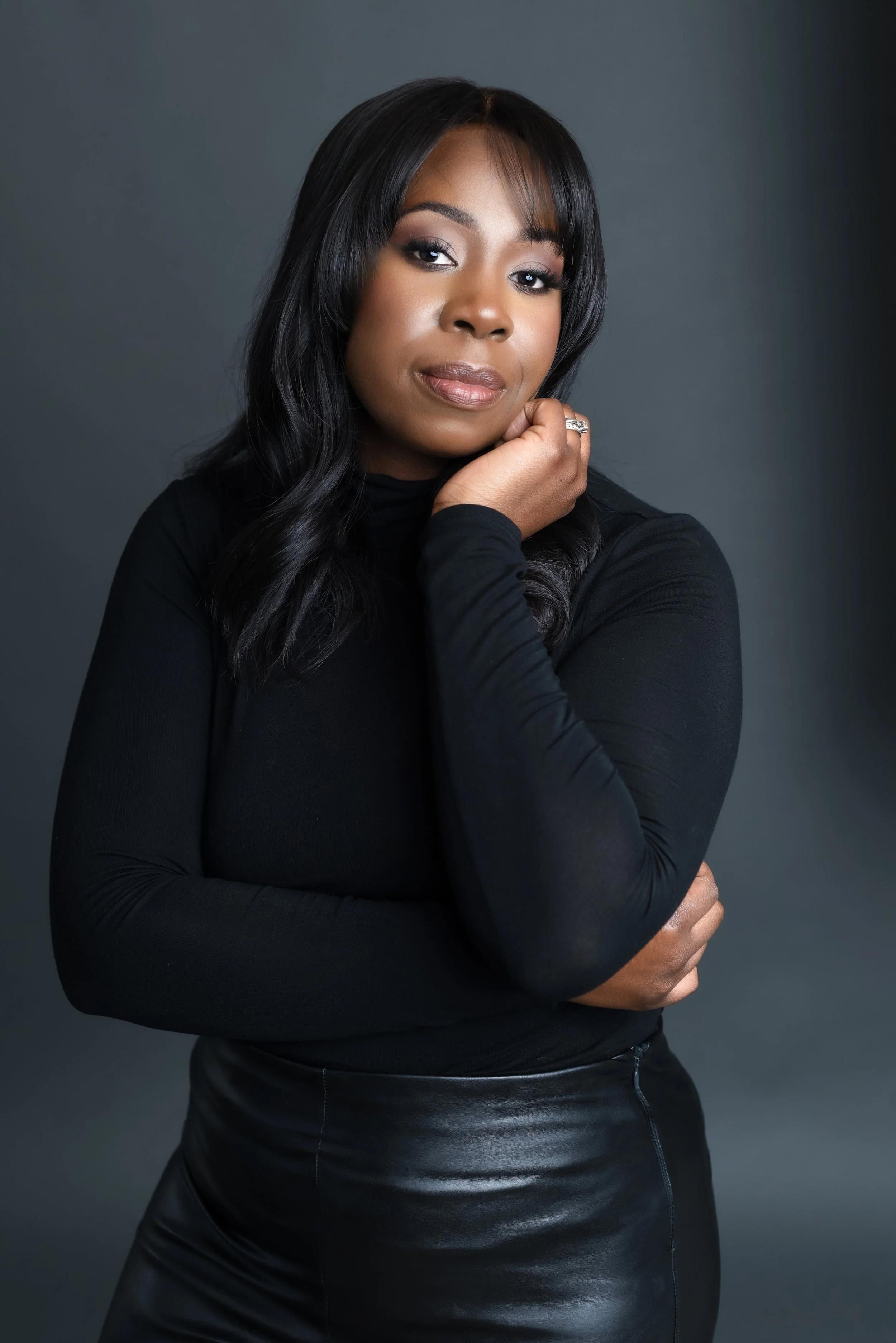 Sabrina Giscômbe with dark hair wearing a black top and leather skirt against a gray background.