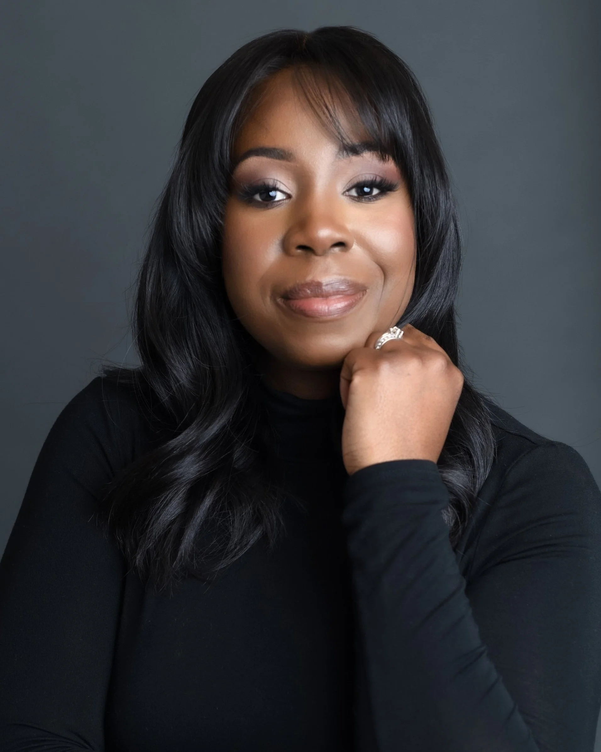 Sabrina Giscômbe  with dark hair, wearing a black top, resting her chin on her hand with a ring, against a plain dark background.
