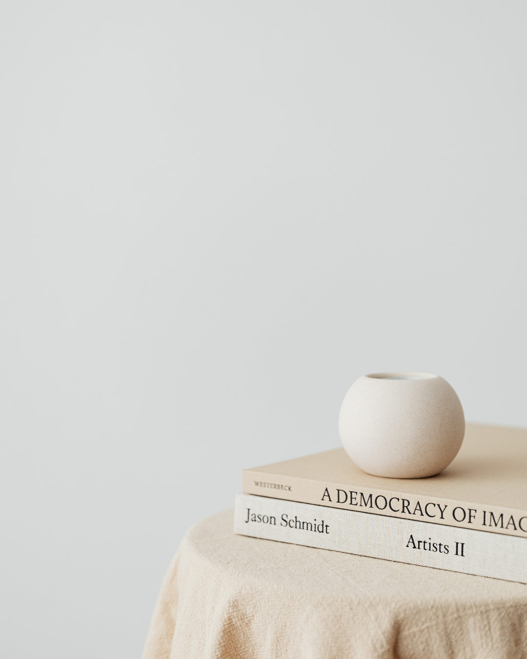 A small, round white ceramic vase on top of two stacked books, one titled 'A Democracy of Imagination' and the other 'Artists II' by Jason Schmidt, placed on a beige cloth-covered surface against a plain, light background.