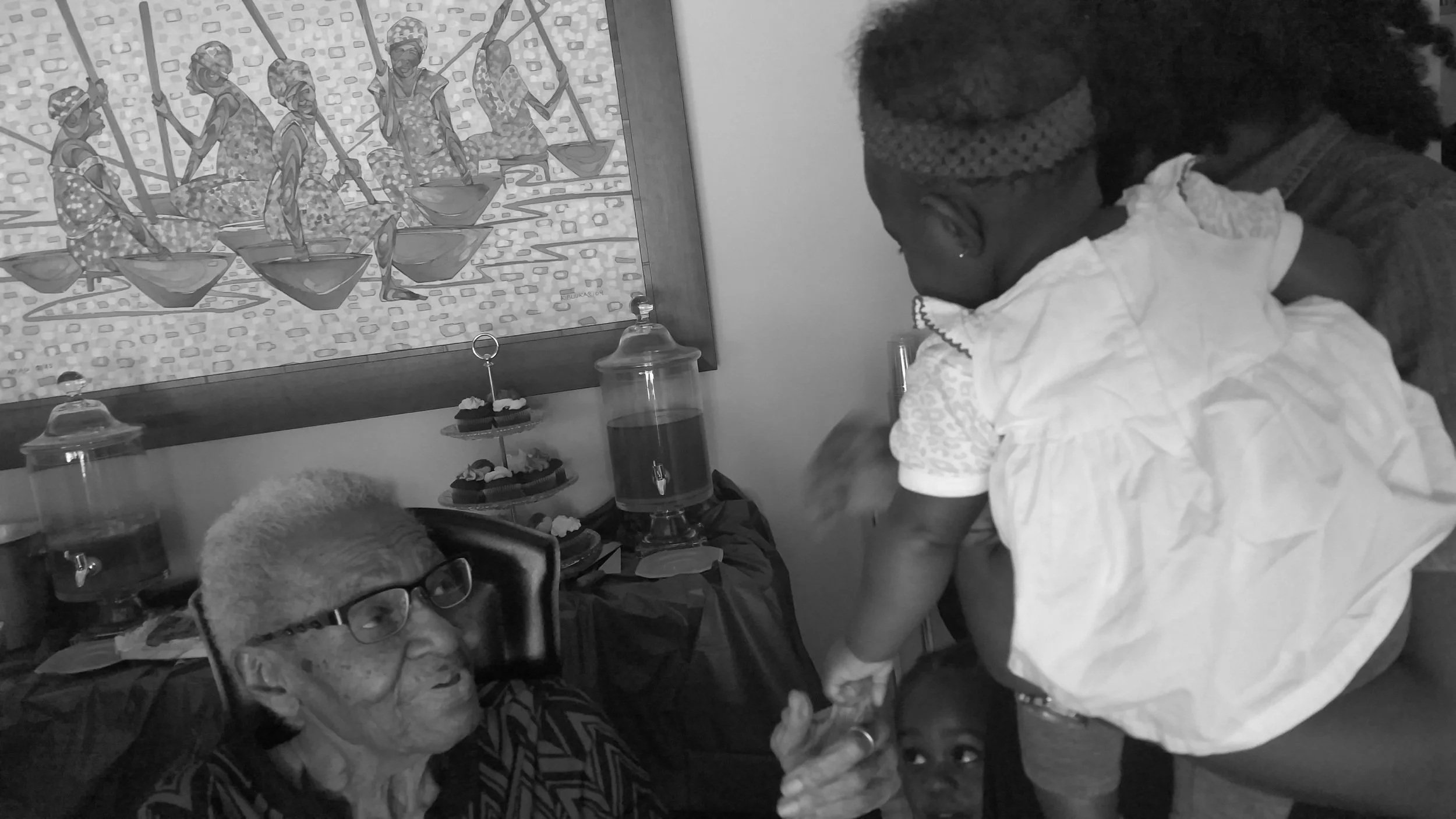 An elderly woman with glasses sitting down and holding hands with a young girl who is being lifted by a person. The scene appears to be at a celebration or gathering.