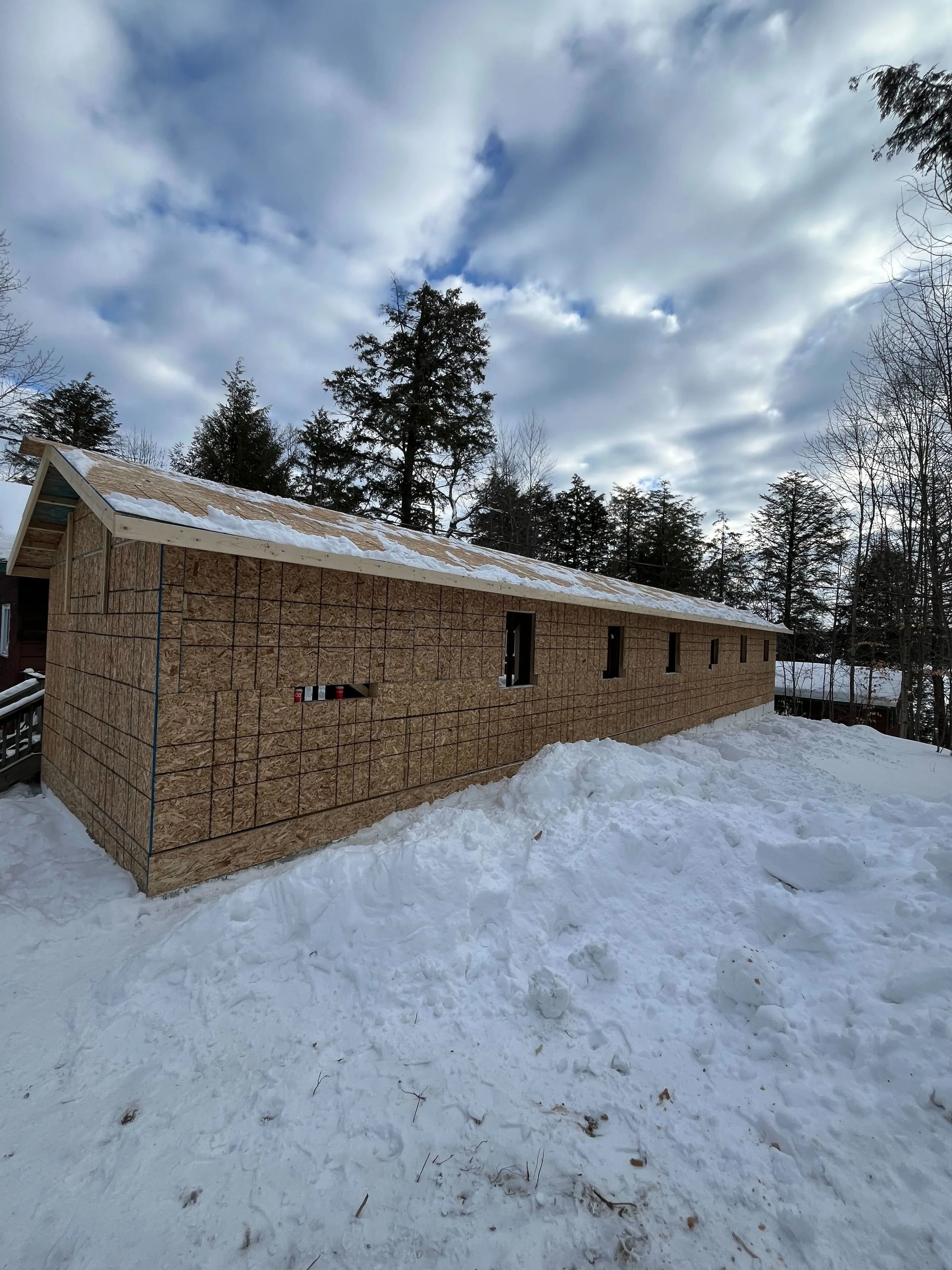 Construction of a house with wooden sheathing on snowy ground, surrounded by trees, with a cloudy sky overhead.