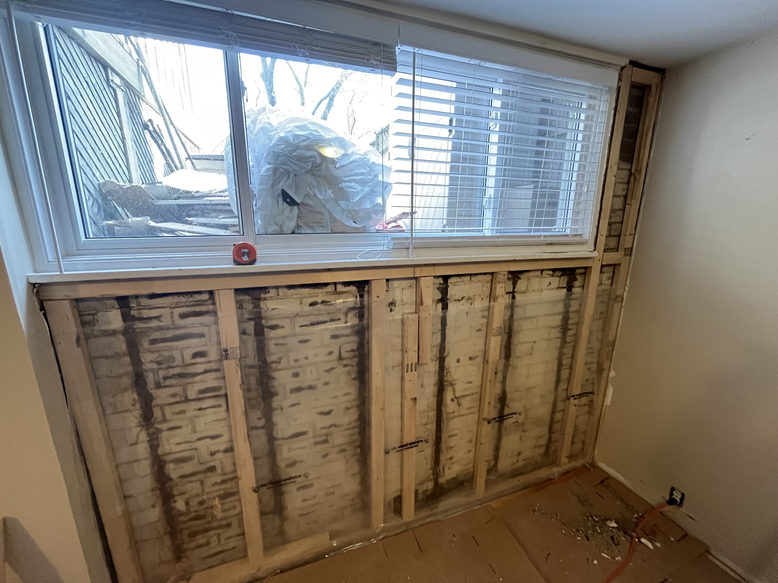 A partially removed wall reveals brick behind wooden framing, with a large window and blinds. Outside, a snow-covered yard and trees are visible, along with a large mass wrapped in white material outside the window.