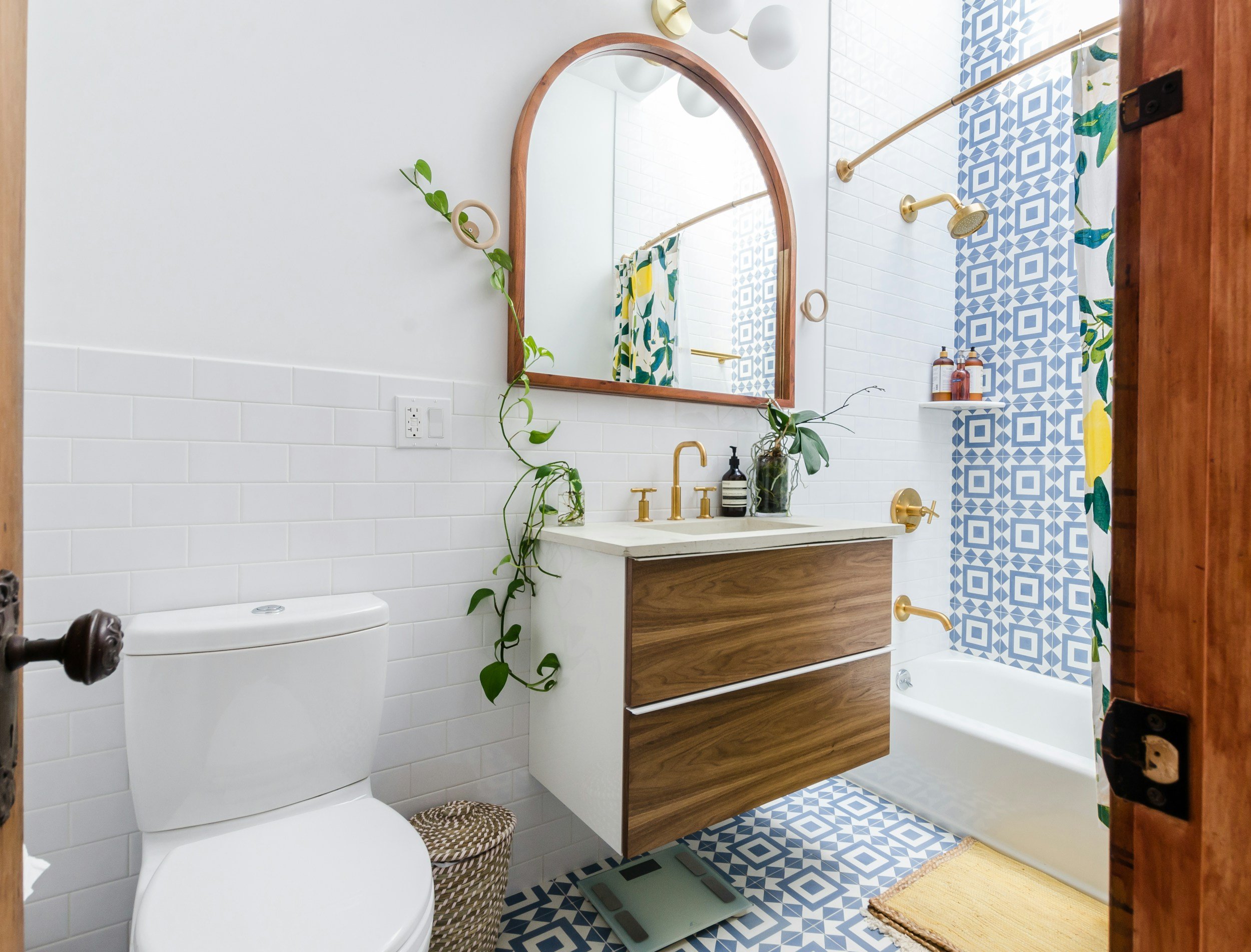 Bathroom with white subway tile halfway up the wall, wooden accents, and blue geometric patterned tiles in the shower and on the floor. Features a white toilet, a floating vanity with a wood front, a leaf-shaped mirror, gold fixtures, and a shower with a gold curtain rod, showerhead, and controls. Decor includes potted plants, bottles, a scale, and a beige bath mat.