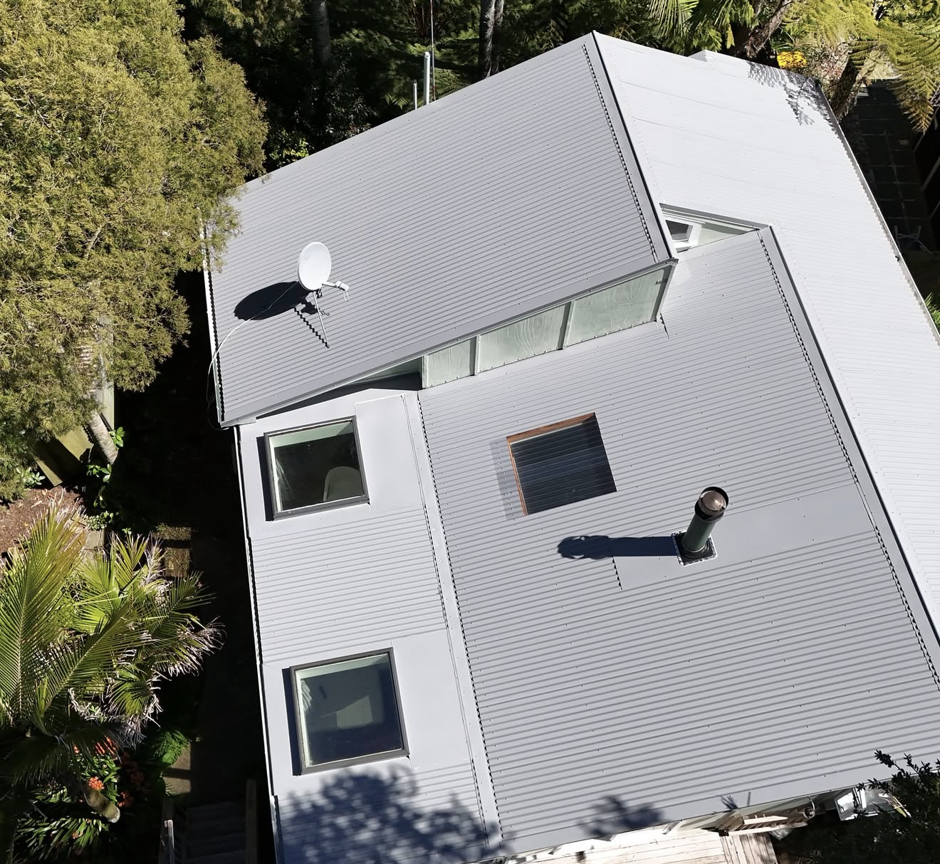 Aerial view of a modern house with a gray corrugated metal roof, several windows, a satellite dish, a chimney, and surrounded by trees and greenery.