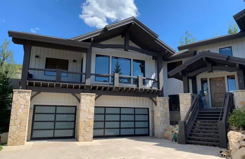 Modern two-story house with large glass windows, stone and wood exterior, and a double garage in the front.