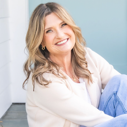 A smiling woman with blonde hair, wearing a light-colored jacket and blue jeans, sitting indoors by a white wall and a blue background.