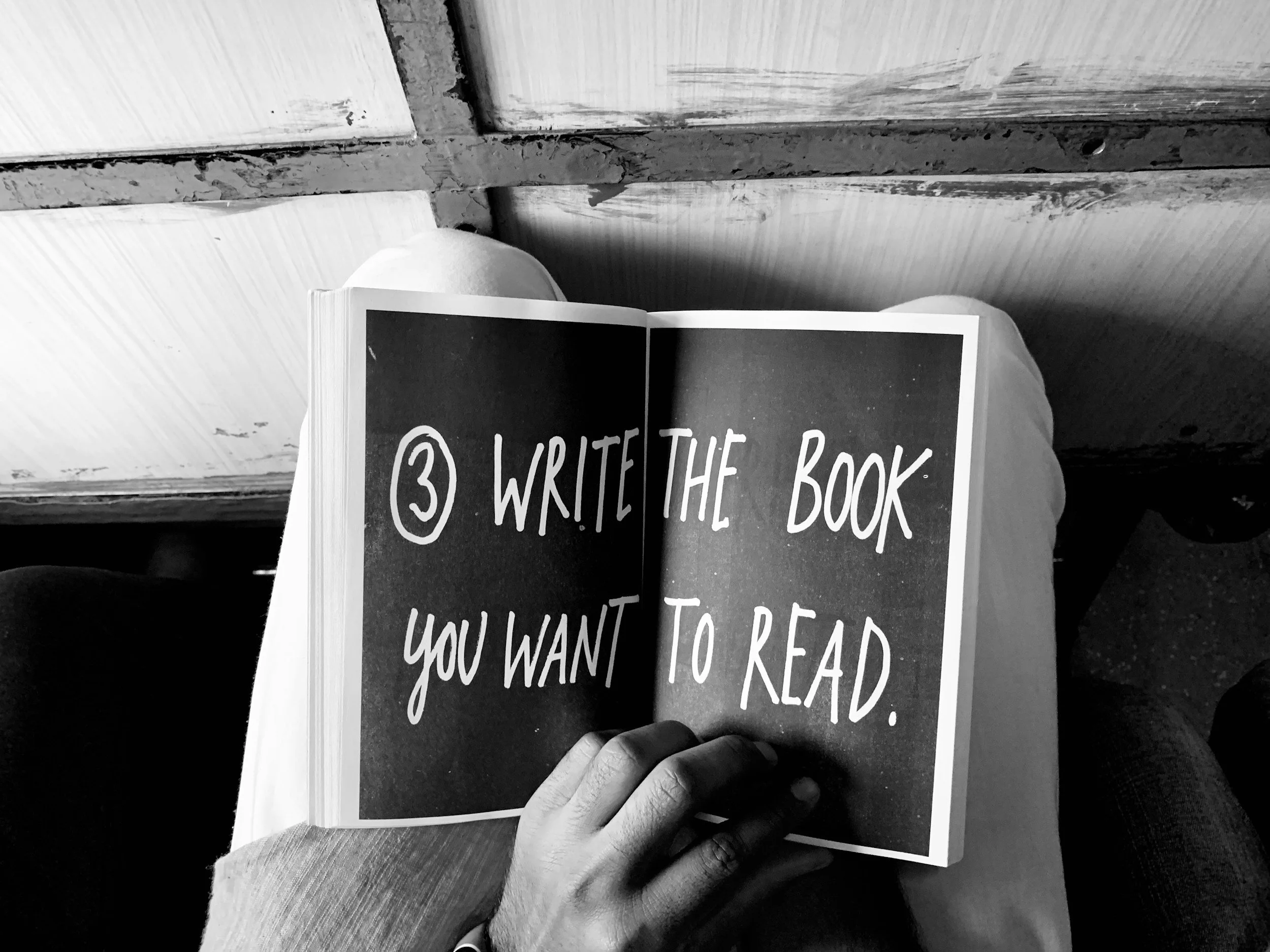 A person is holding an open book with a handwritten message on black pages that says, '3. Write the book you want to read.' The person's hand is visible, and they are wearing a wristwatch. The setting appears to be outdoors or in a casual environment.