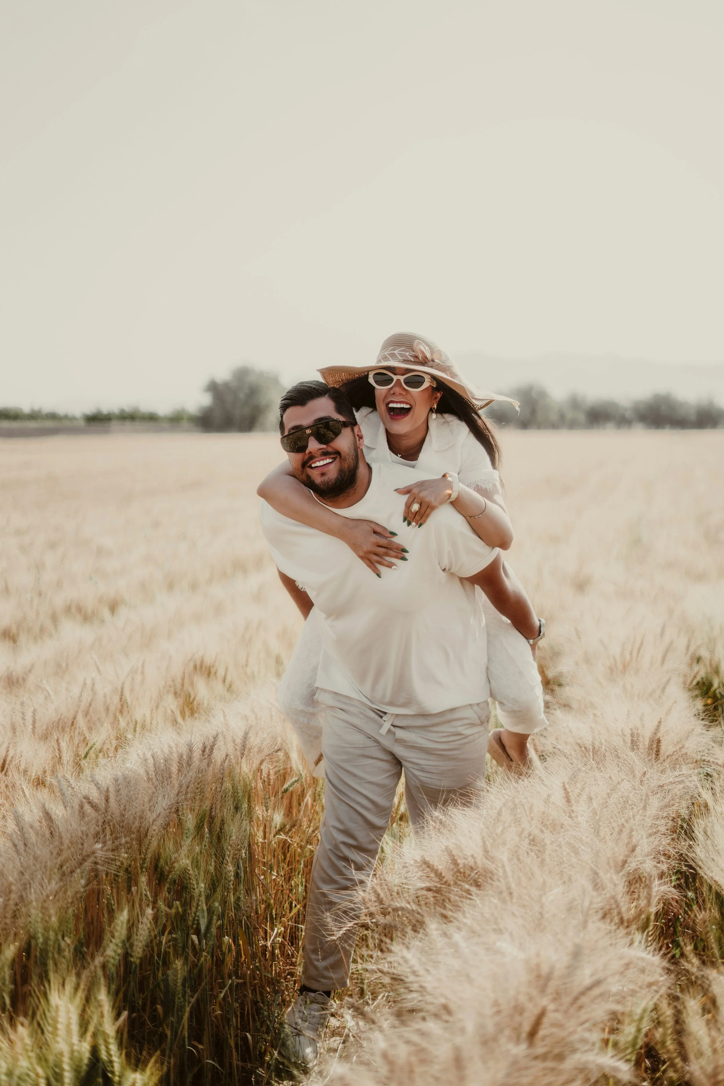 A happy couple enjoying a piggyback ride in a golden wheat field, both wearing sunglasses and smiling after premarital counseling in Orange County.