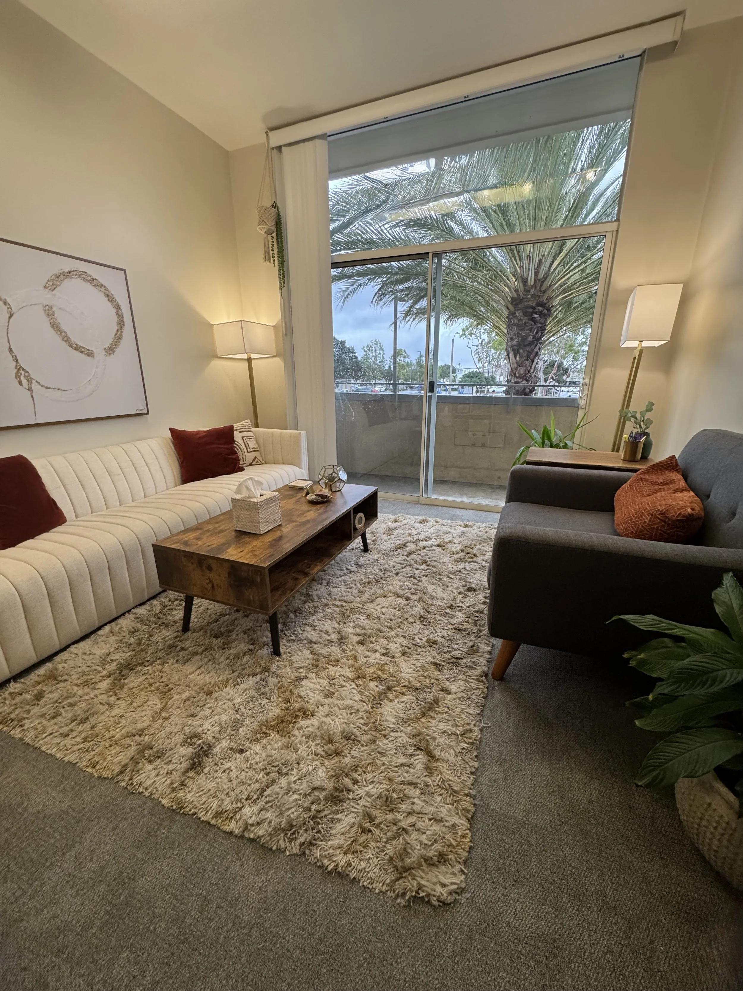 Living room with a beige tufted sofa, a dark gray armchair, a wooden coffee table, a fluffy area rug, floor lamps, and a sliding glass door leading to a balcony with a view of a palm tree and outdoor area.